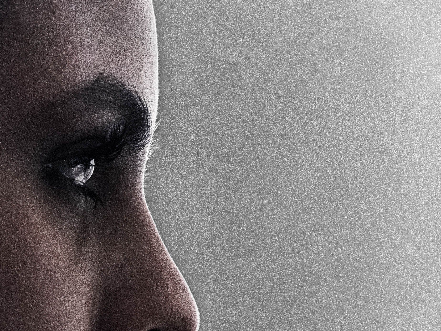 Extreme close-up of a woman's eye and nose in profile, heavy grain texture, soft light tracing one side of the face against a plain background