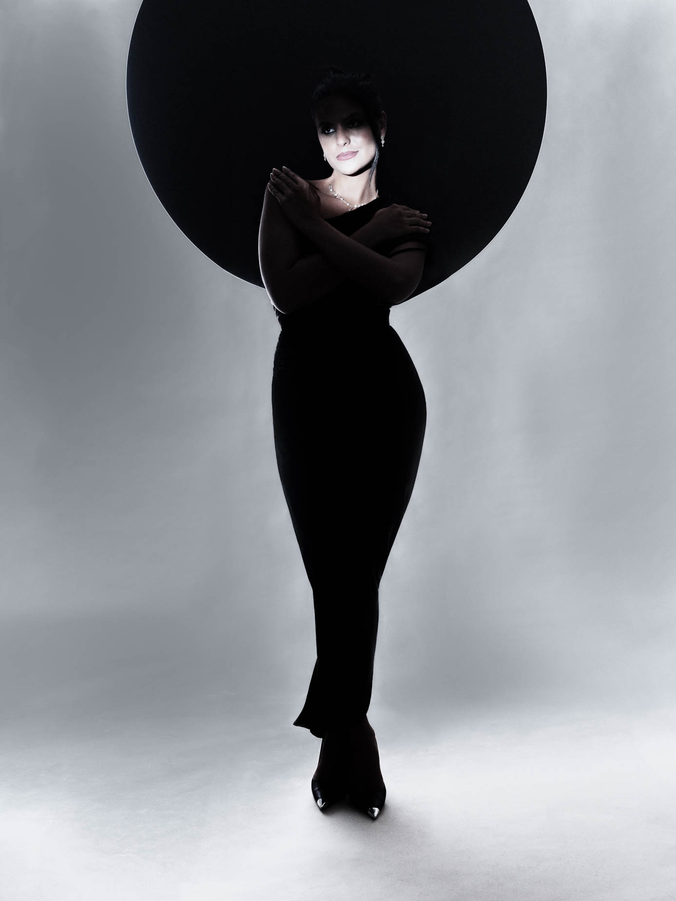 Woman standing beside a large dark circular prop, wearing a one-shoulder black dress with necklace, soft light on the face