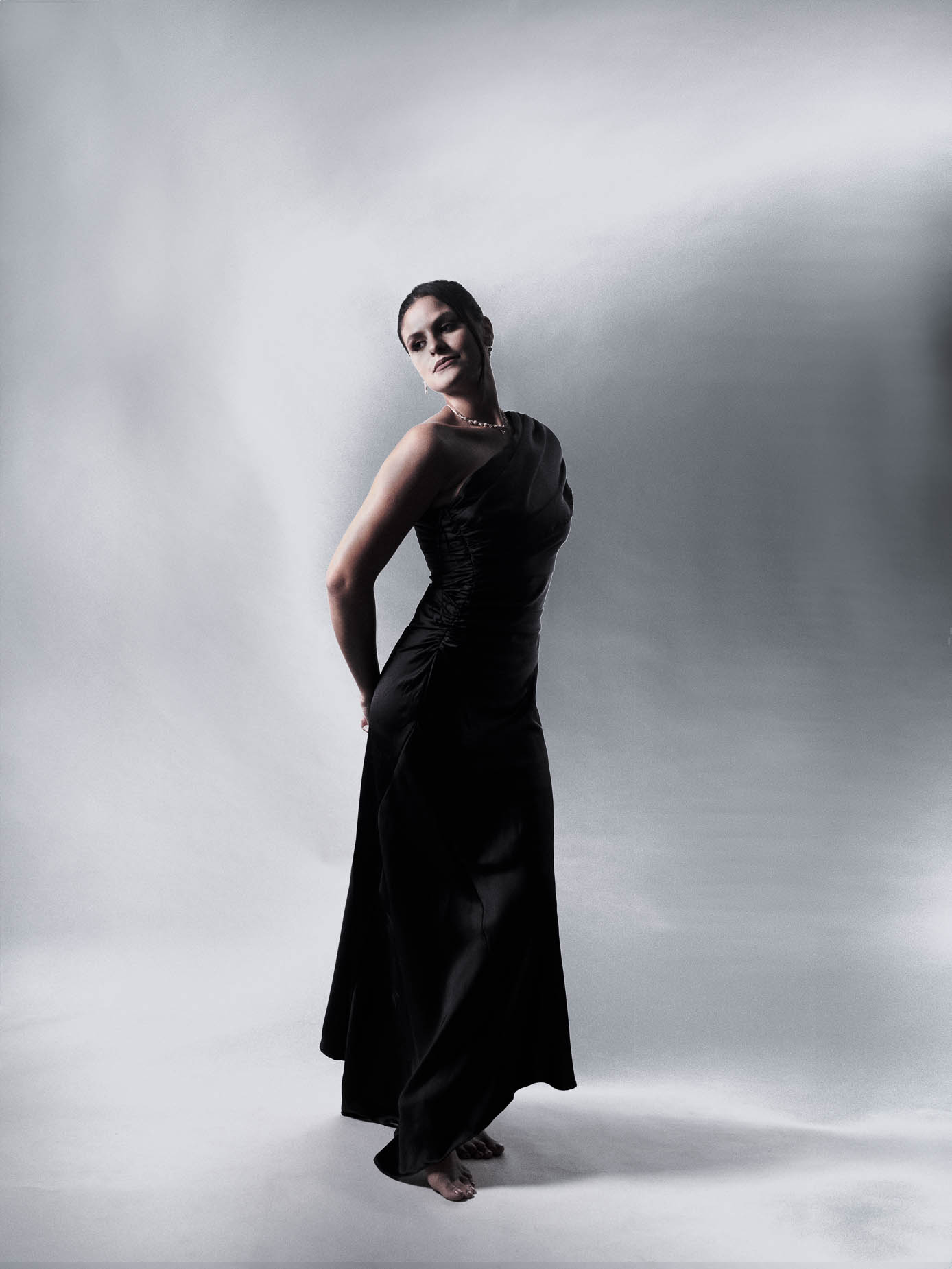 Woman standing full length in a black one-shoulder gown, one hand on her hip, looking back over her shoulder