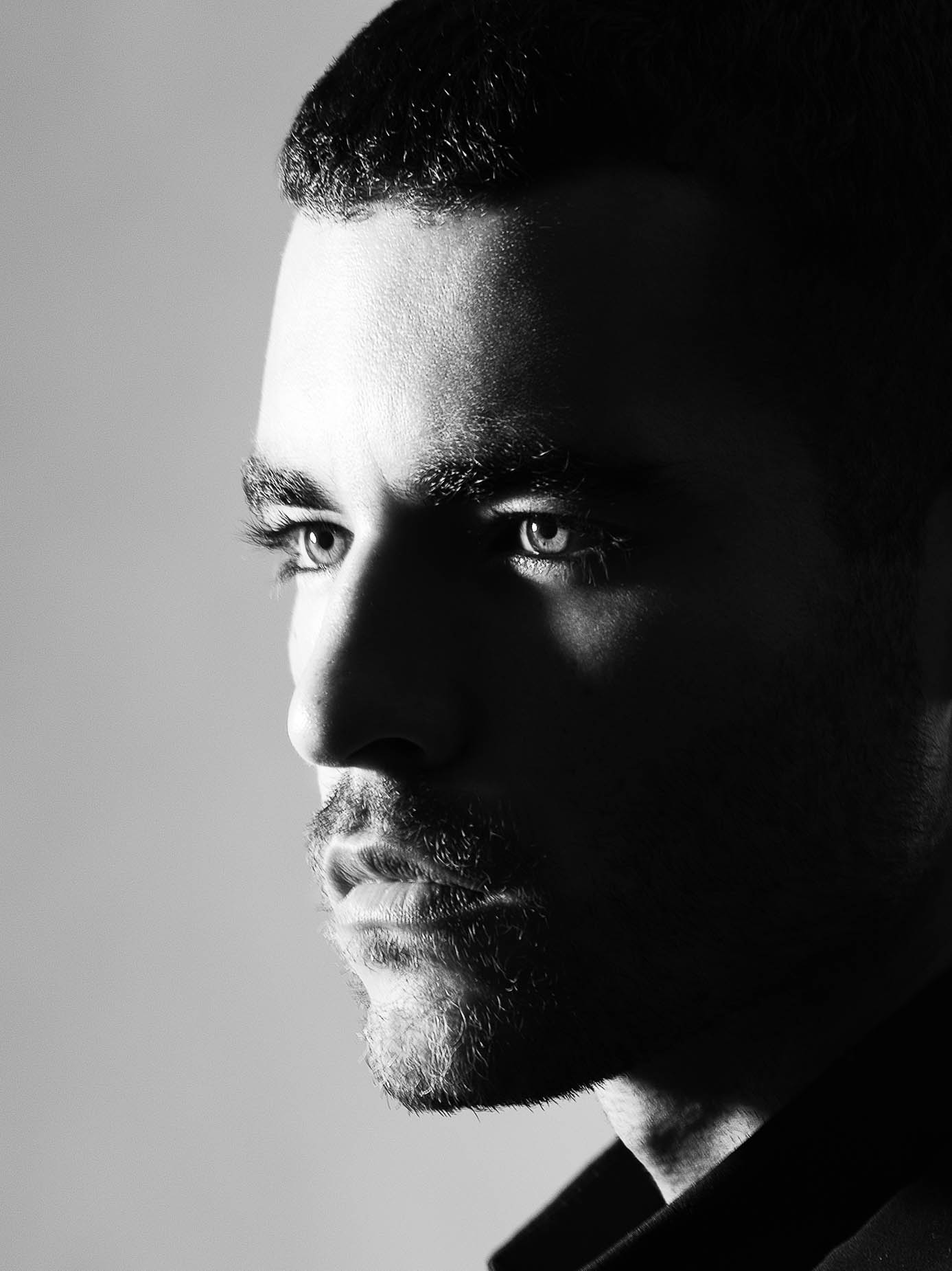 Extreme close-up of a man's face lit sharply from one side, a hard shadow splitting the features, eyes catching the light against a plain grey background