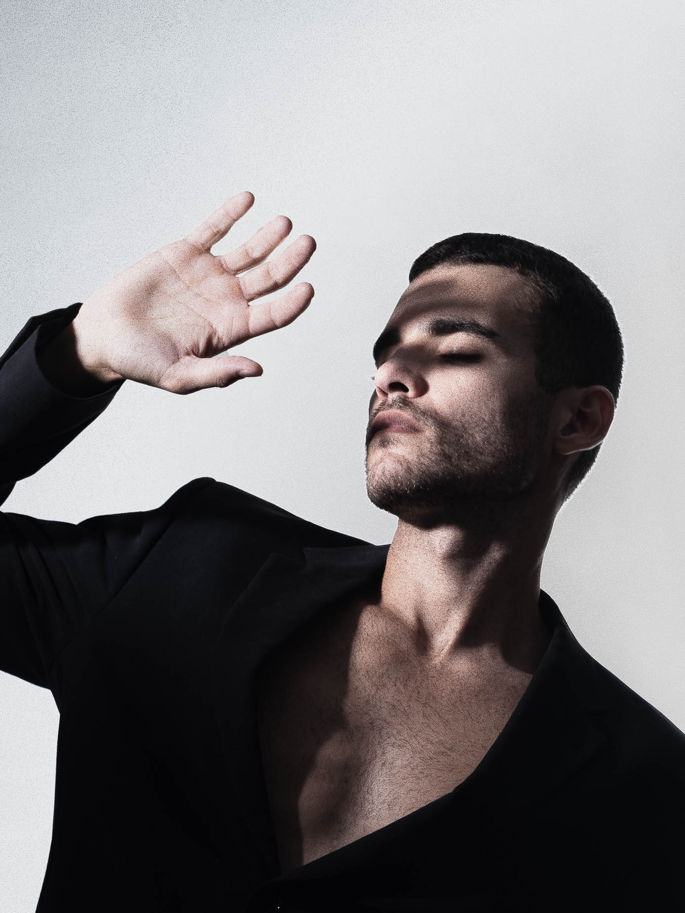 Man in an open black wrap outfit with bare chest, eyes closed, one hand raised in a dynamic dancer-like pose