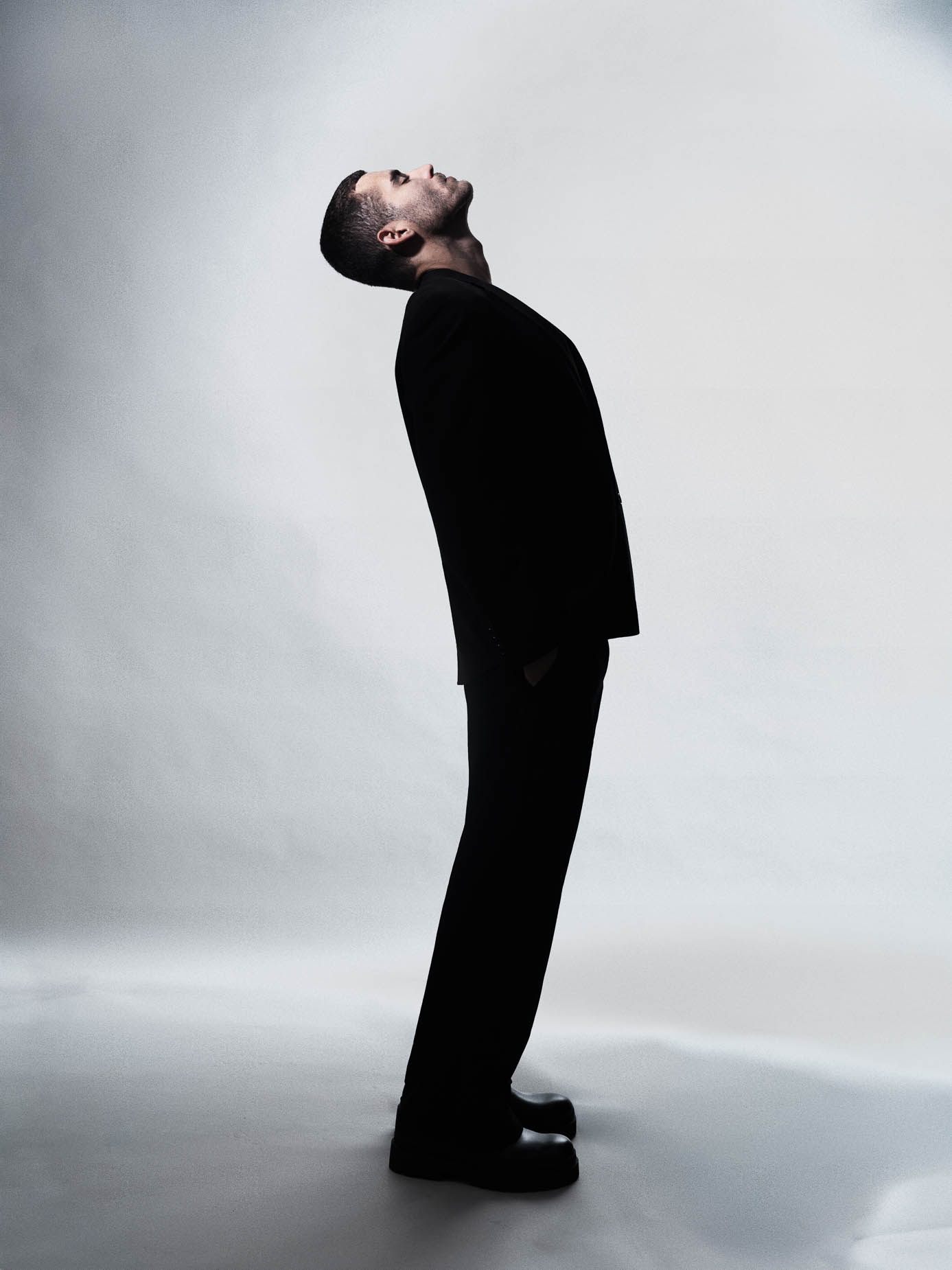 Man standing full length in an oversized black suit, one hand raised, chin lifted, dramatic studio lighting