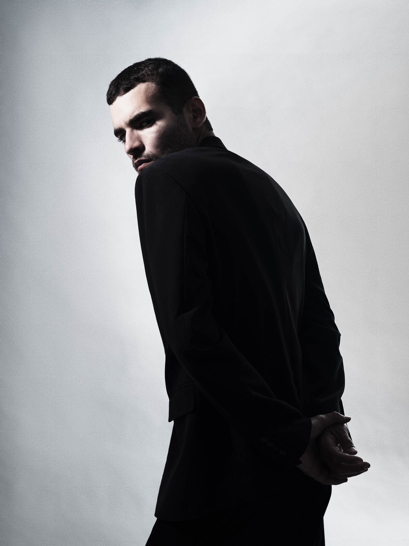 Man glancing back over his shoulder in a black jacket, hands clasped behind his back, moody side lighting