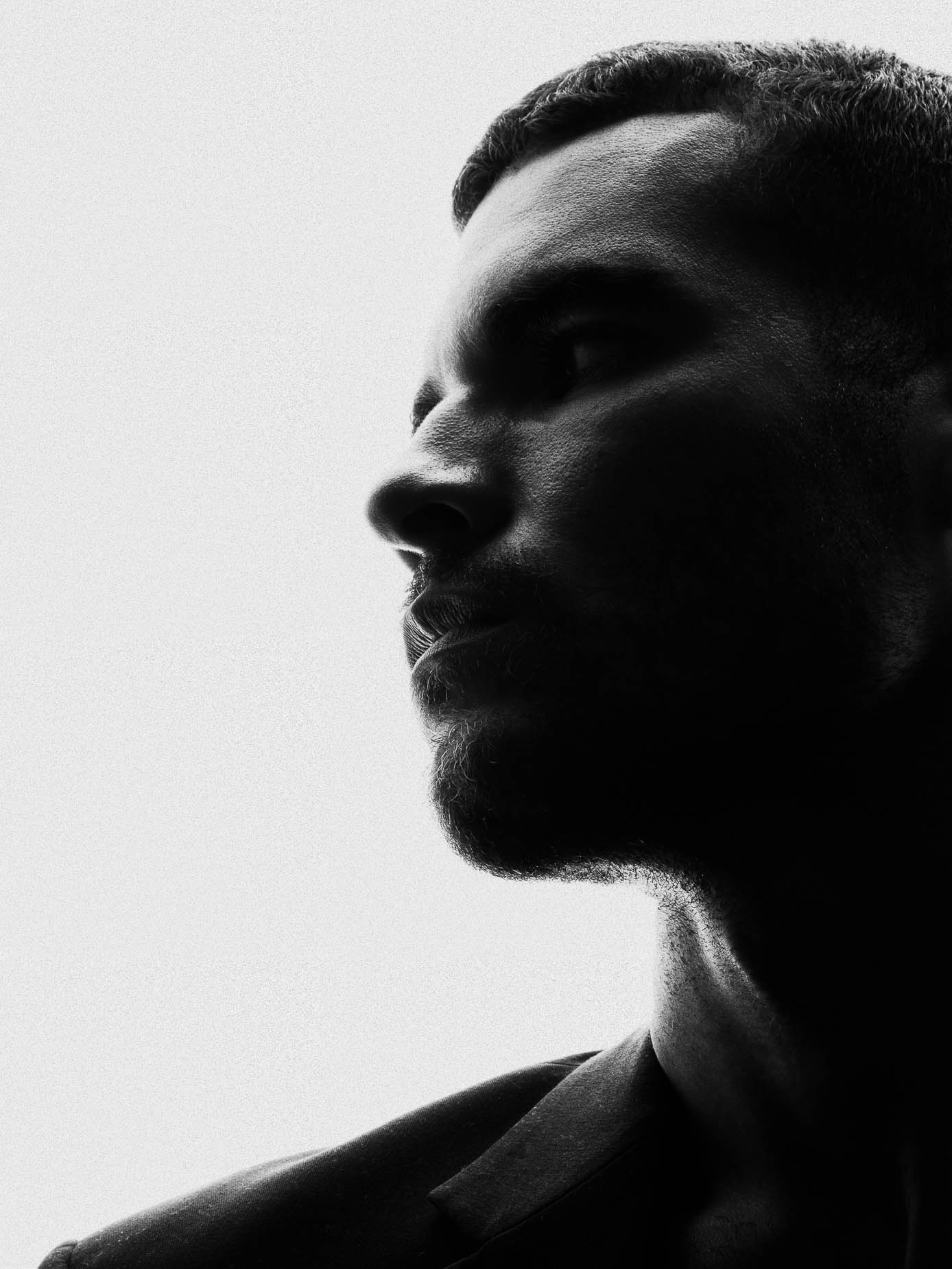 Man in near silhouette, his dark jacket collar forming a sharp graphic V-shape across the shoulders, stark high-contrast studio lighting