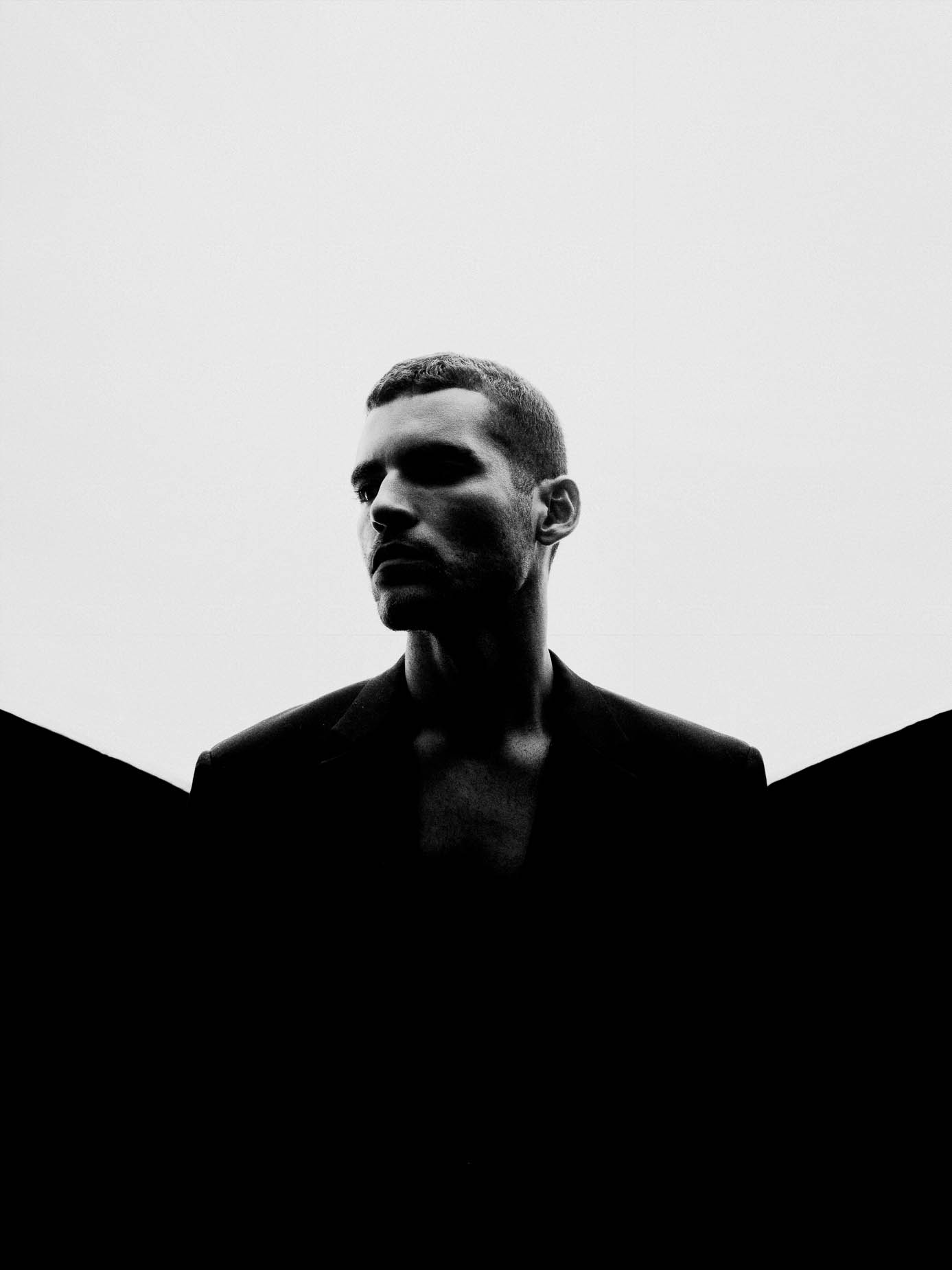 Man in a dark blazer with eyes closed, chin lowered, soft studio light against a plain background