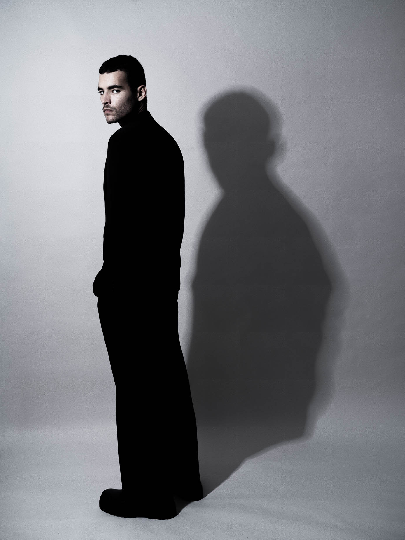 Man seen from the side in black clothing, head turned toward the camera, a large dramatic shadow of the silhouette cast on the wall behind