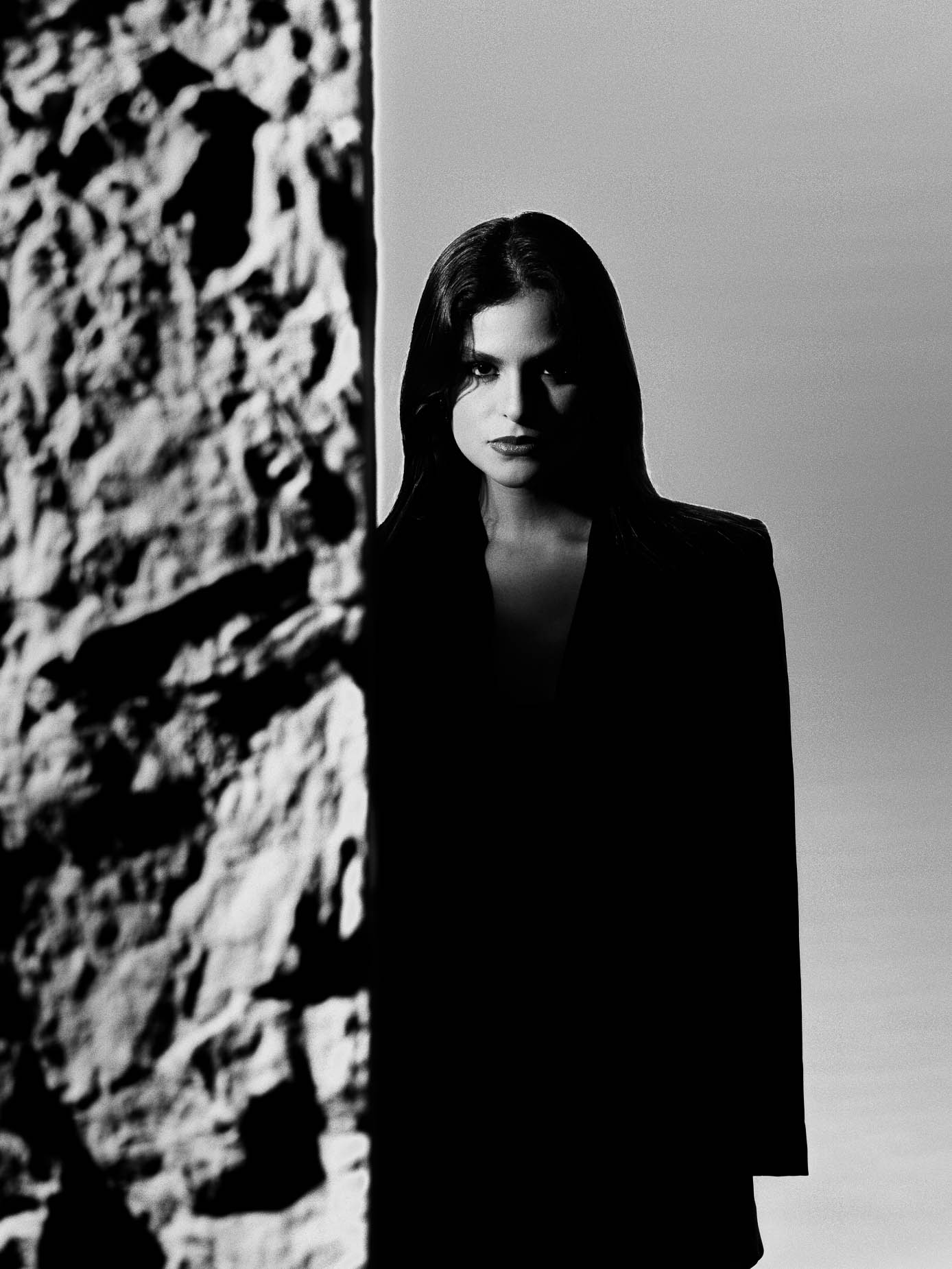 Woman in a black dress stepping out from behind a rough stone pillar, dark lipstick, direct gaze at the camera