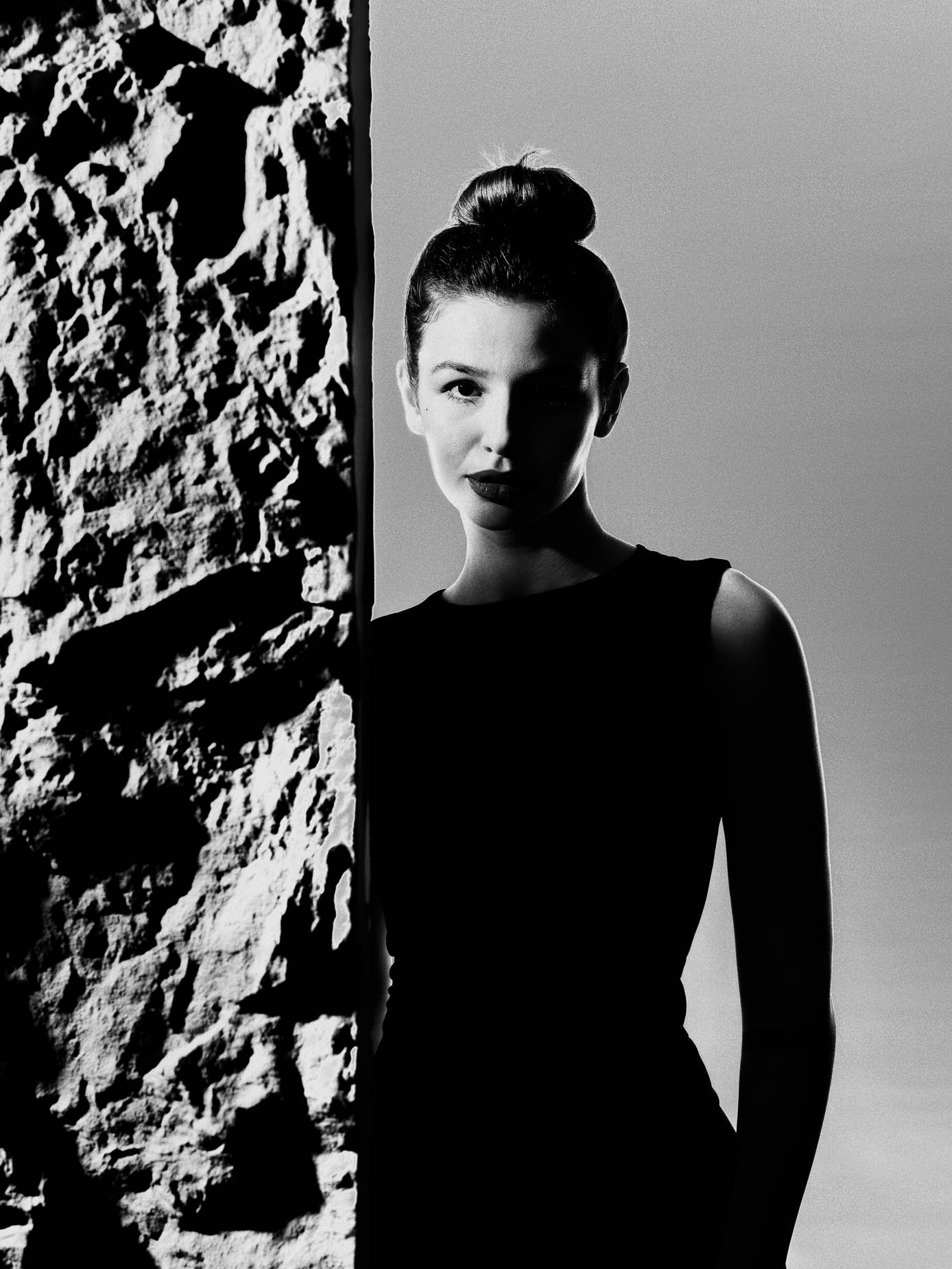Woman with hair in a bun stepping out from behind a rough stone pillar, wearing a black dress and dark lipstick, direct gaze at the camera