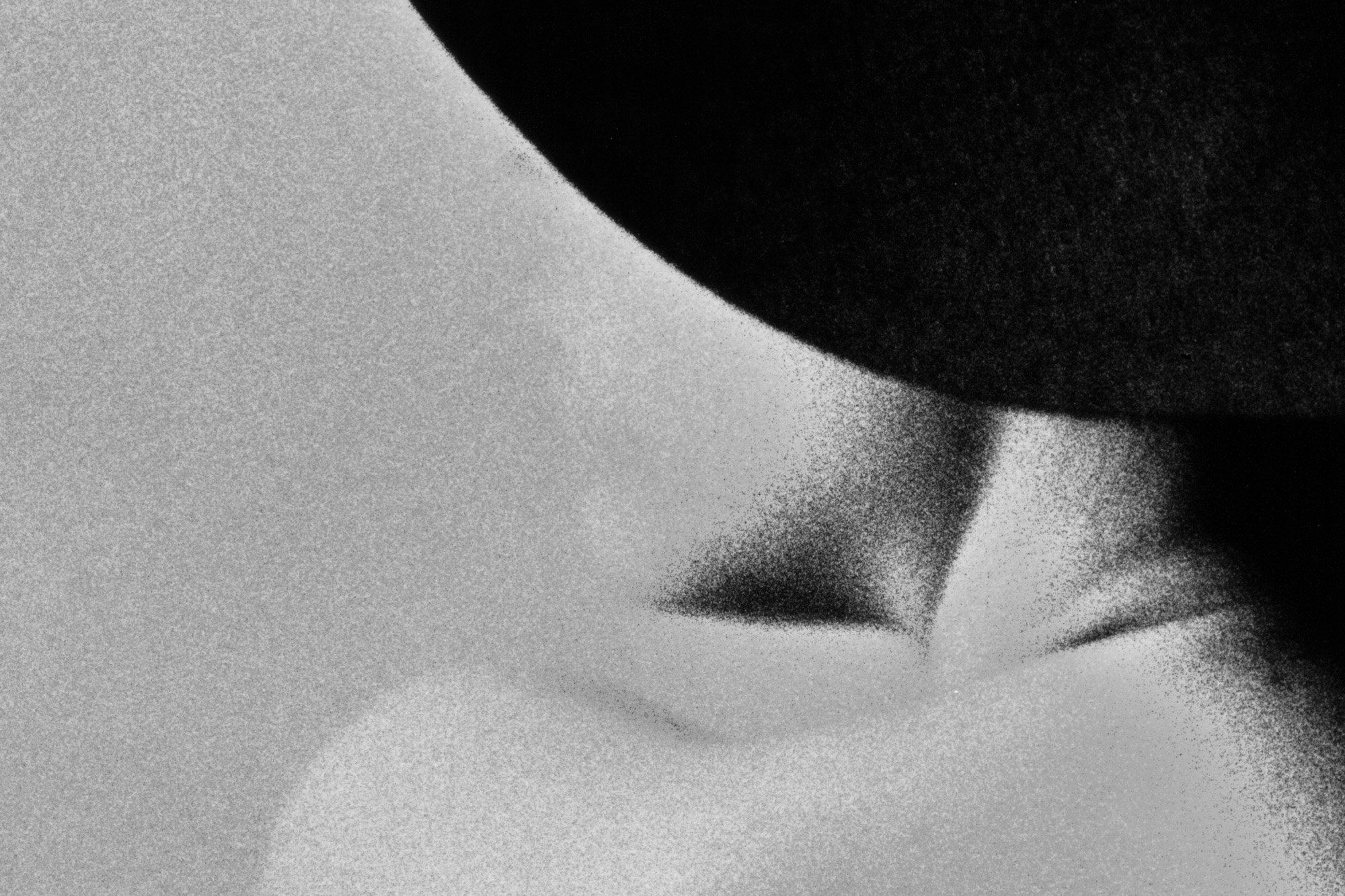 Extreme grainy close-up of a bare shoulder and neck curving beneath a dark rounded shape, high-contrast black and white texture with heavy visible film grain