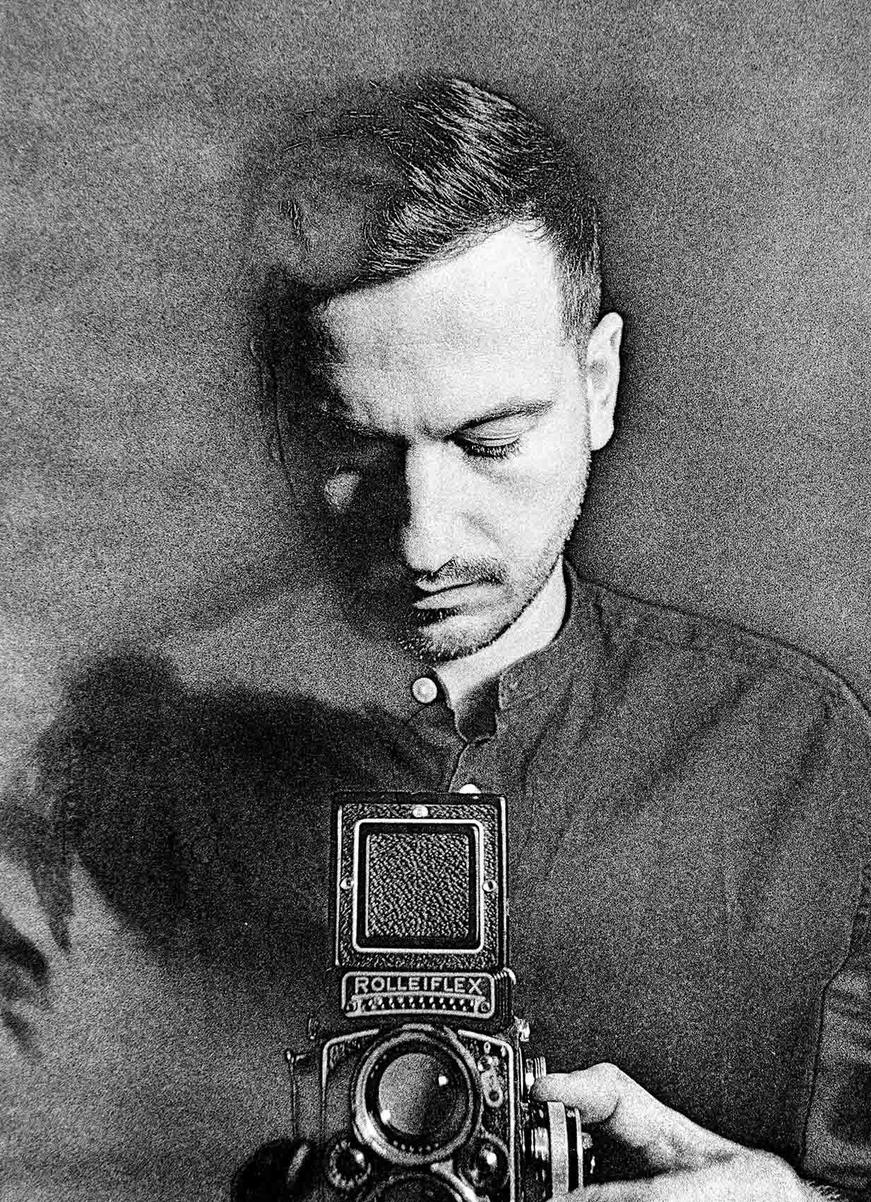 Black and white self-portrait of Hugo Chazot-Ranquet looking down at a Rolleiflex twin-lens camera held at chest height, grainy film texture, soft shadow cast on the wall behind him