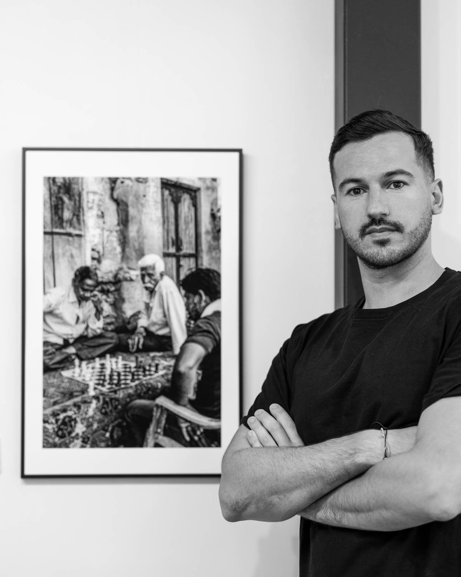 Hugo Chazot-Ranquet standing with arms crossed beside a large framed black and white street photograph of three men playing chess in a weathered alleyway
