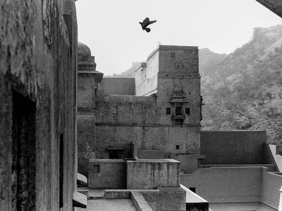 A bird flies above the weathered stone towers of a hillside fort, with hazy mountains and more fortress walls visible in the distance