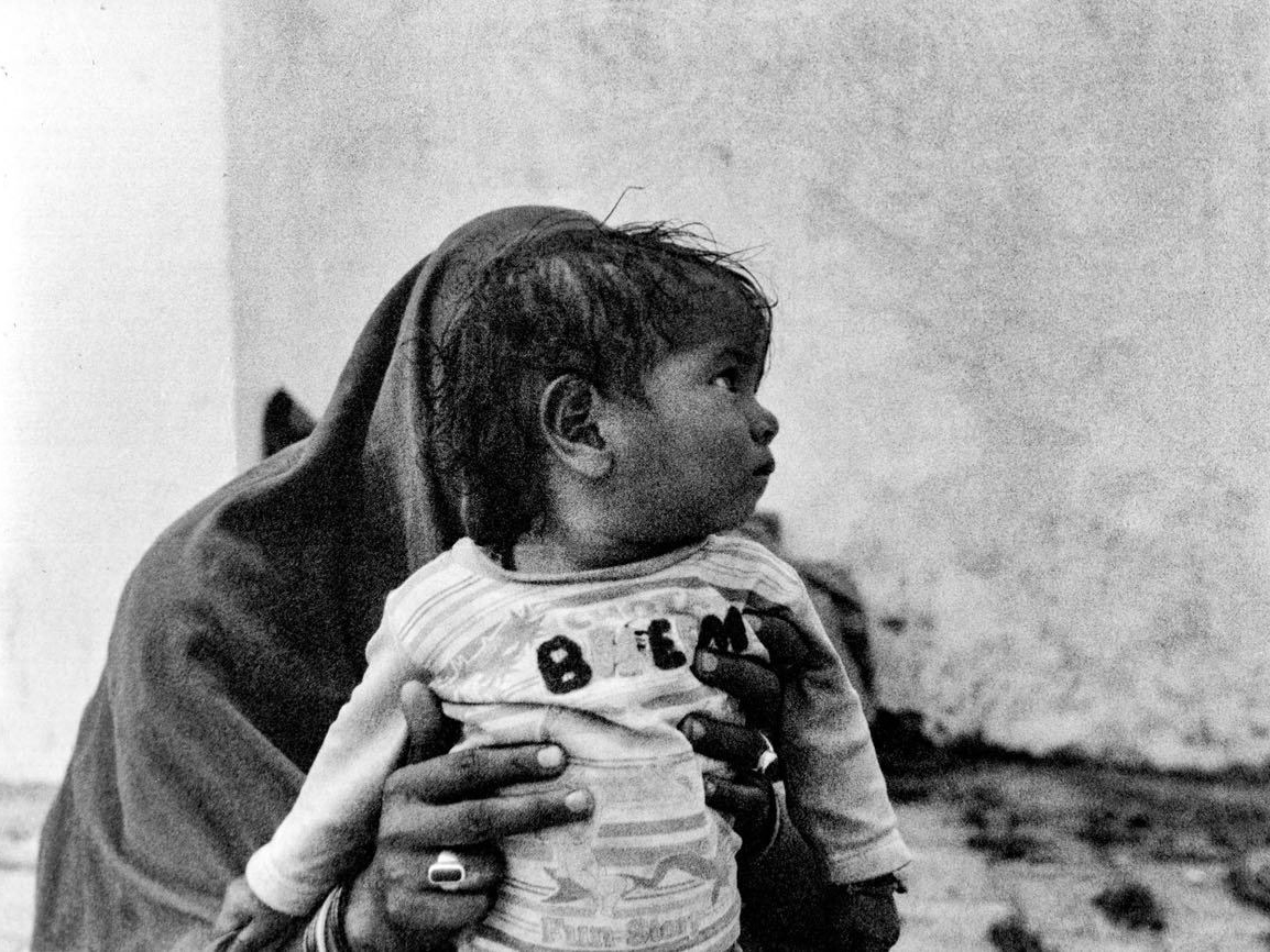 A woman wrapped in a shawl holds a young child close to her chest, the child looking off to the side, against a plain plastered wall
