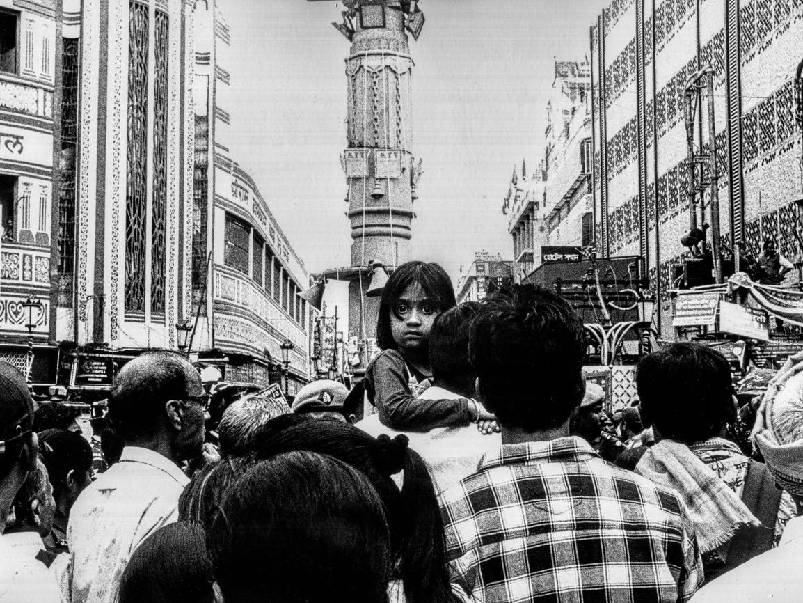 A young girl looks directly at the camera from atop a man's shoulders, rising above a dense crowd in a city street lined with ornate buildings and a tall tower