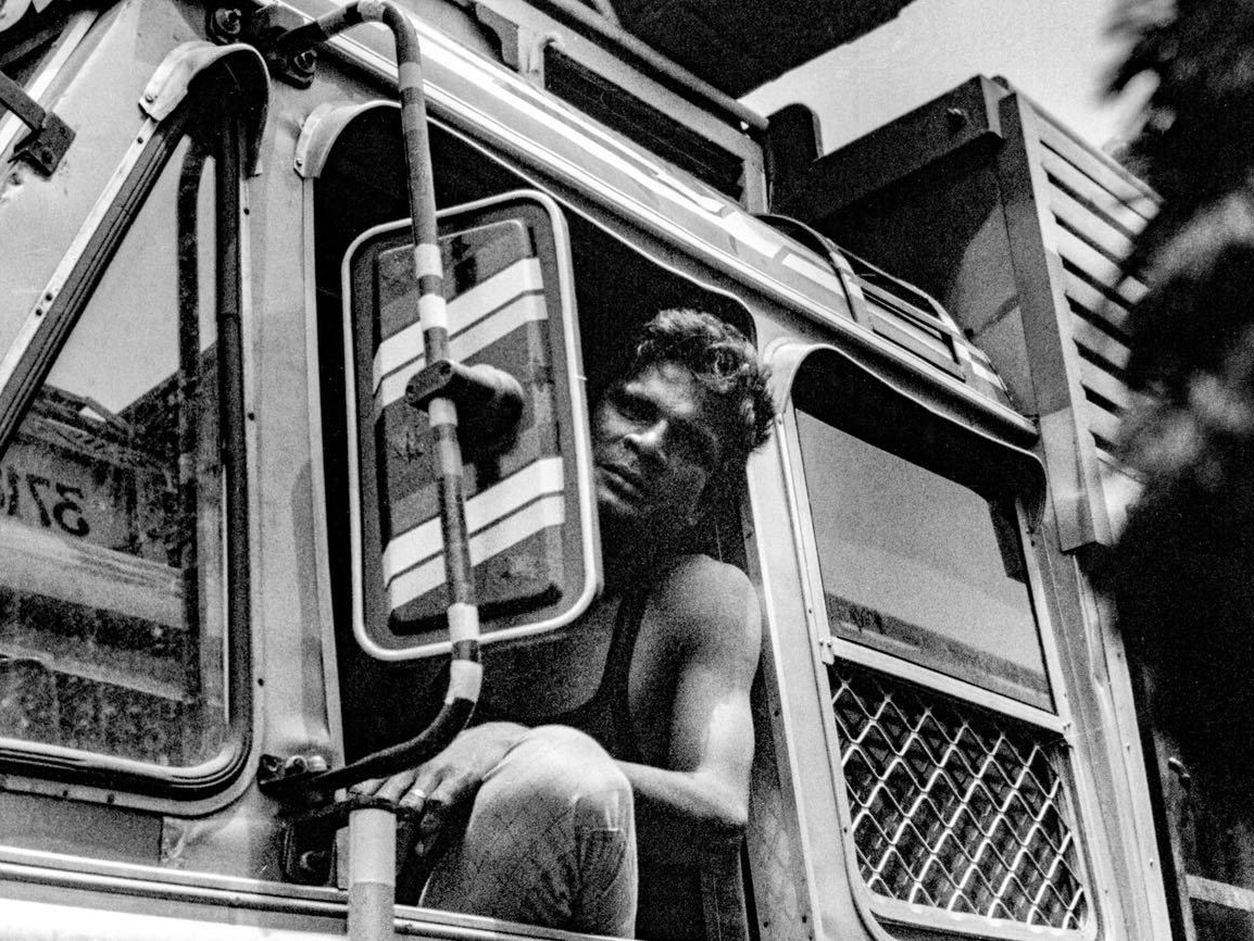 A shirtless man leans out of an open bus window, resting against the frame beside a cracked side mirror, looking down toward the camera