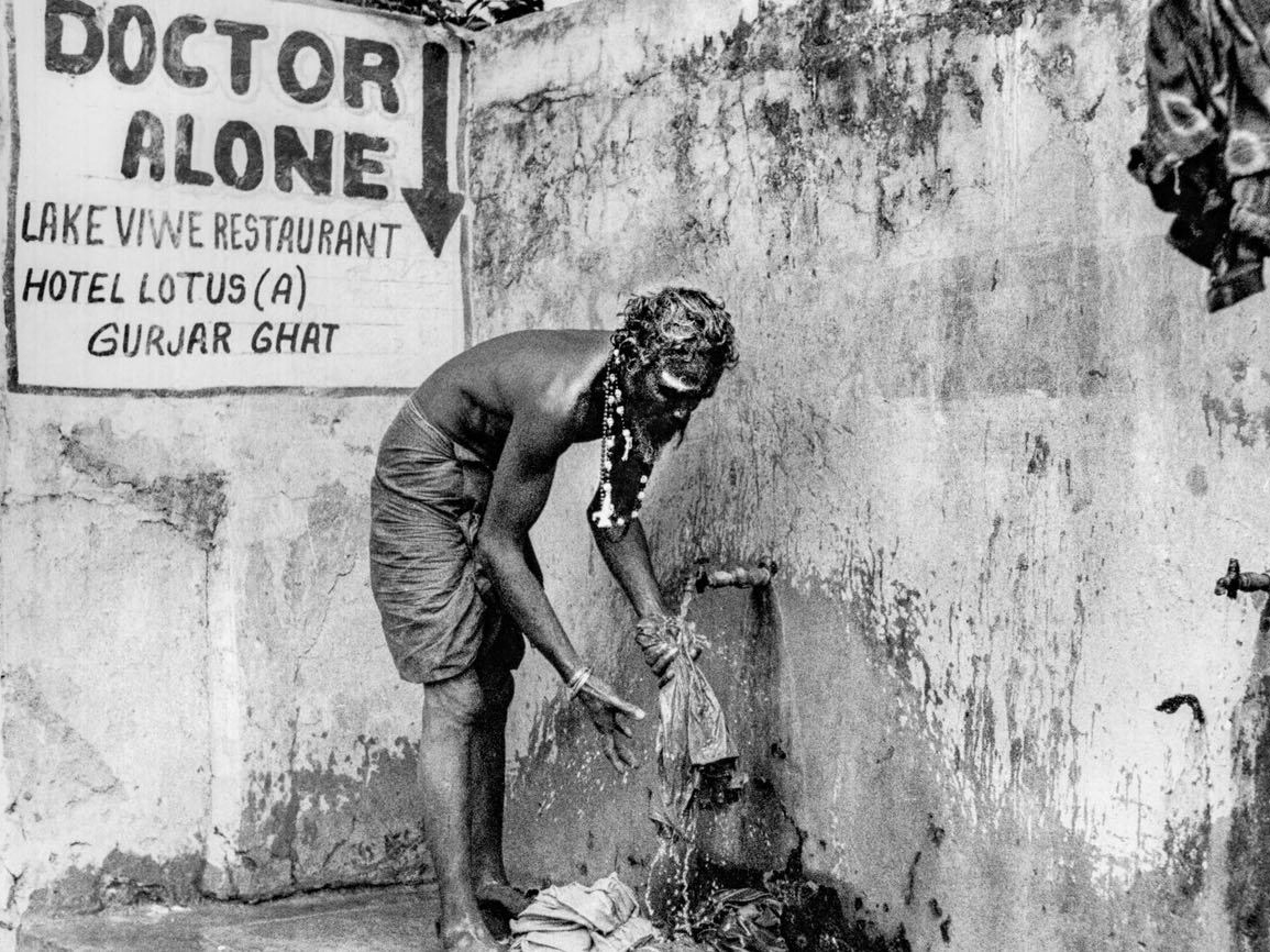 A shirtless man bends over an outdoor tap to wash cloth, wearing a beaded necklace, beneath a hand-painted sign reading Doctor Alone, Lake View Restaurant, Hotel Lotus, Gurjar Ghat