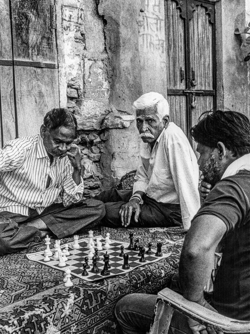 Three men sit on a patterned rug in a stone doorway playing chess, one watching the camera