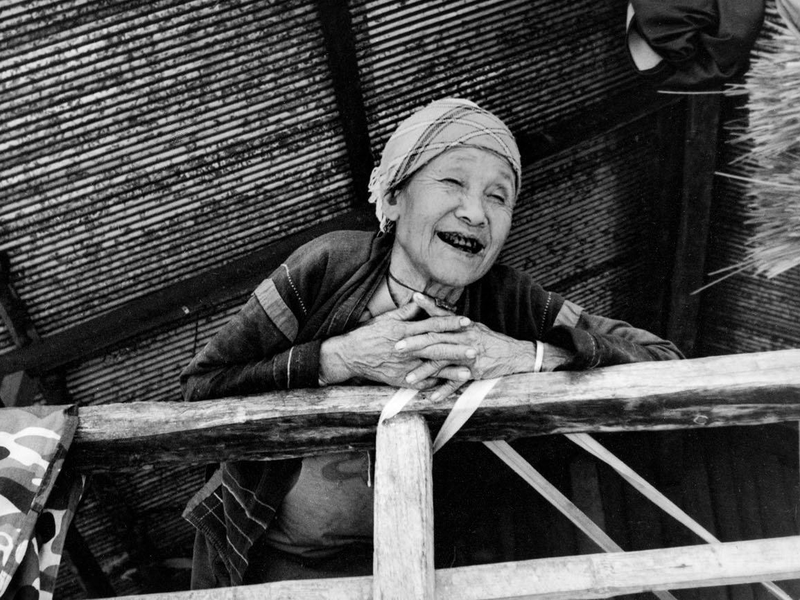 An elderly woman in a headscarf leans on a wooden railing laughing, hands clasped together, standing under a corrugated metal roof