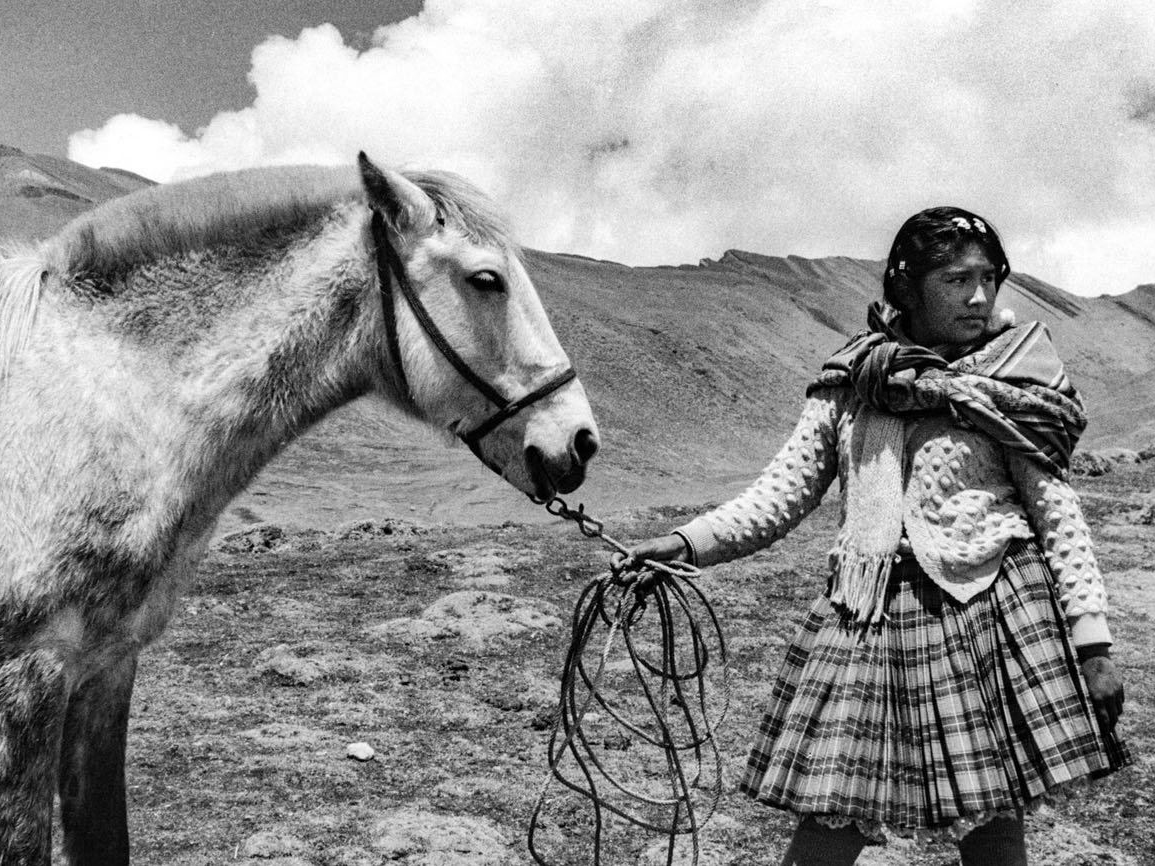A young woman in a plaid skirt and knitted shawl leads a pale horse by its rope, standing on a barren mountainside beneath a large cloud