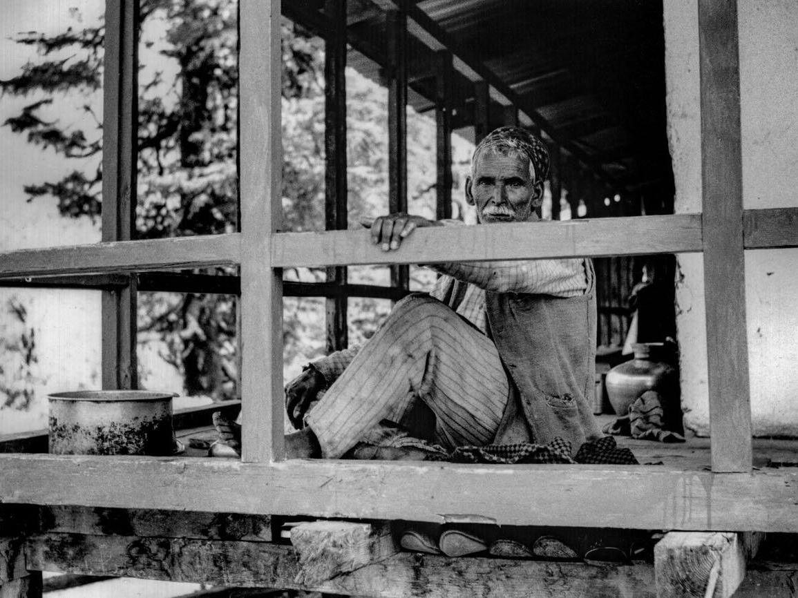 An elderly man with a moustache sits cross-legged on a wooden porch, framed by timber beams and railings, looking toward the camera with a hand resting on the rail