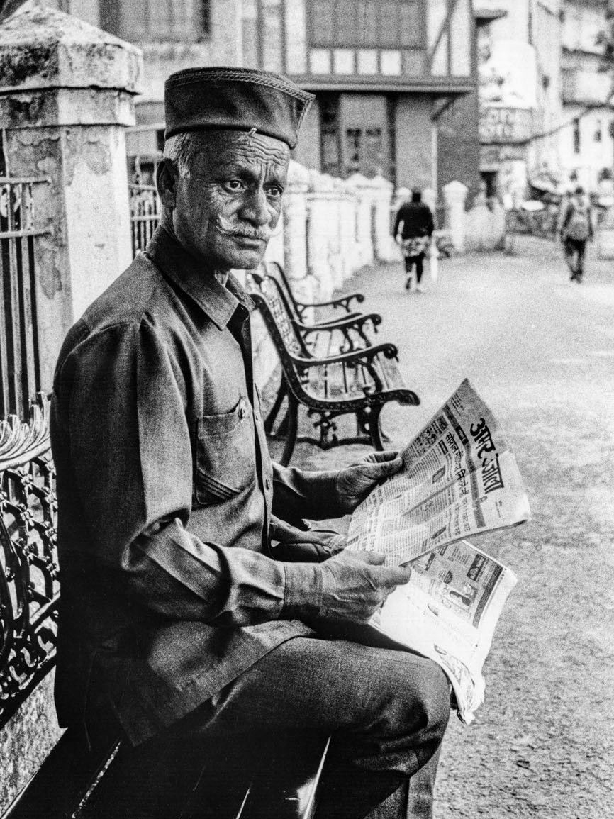 A man in a peaked cap sits on a bench reading a folded newspaper, glancing toward the camera