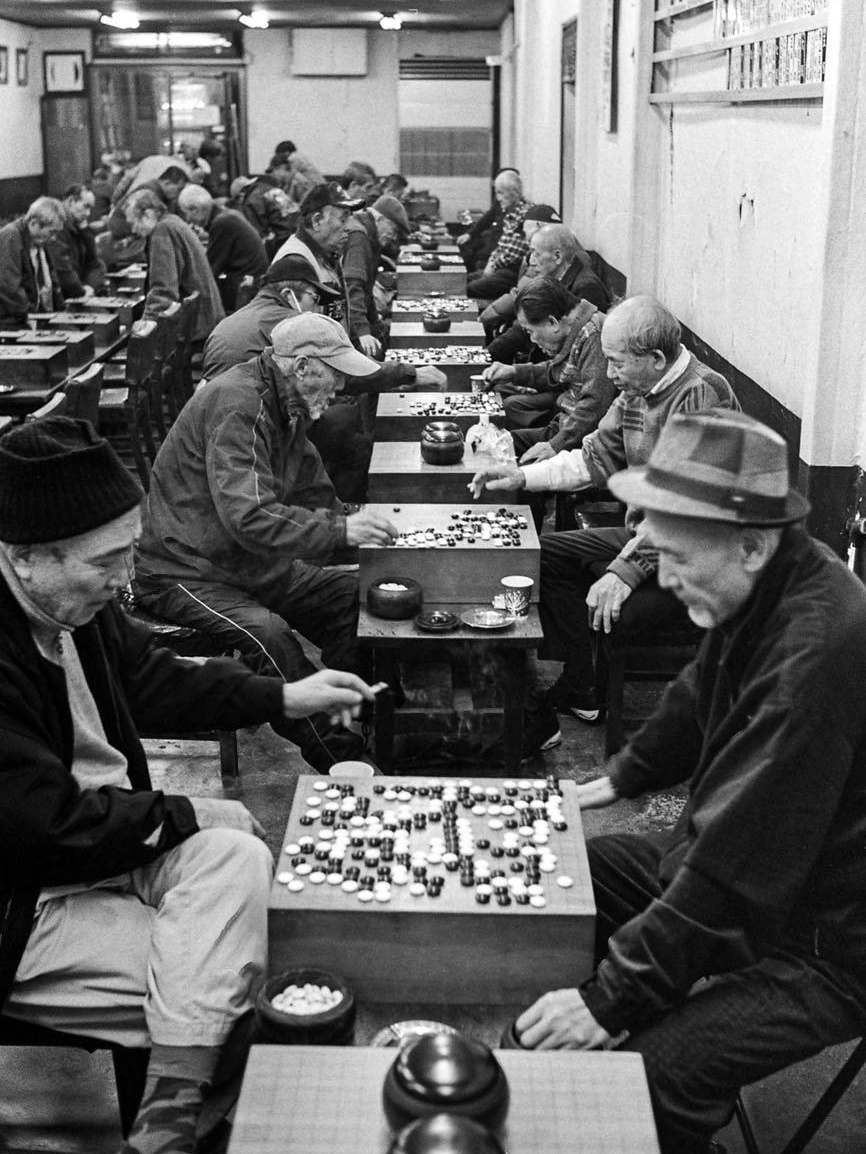 Rows of elderly men play board games at small tables lining a narrow indoor hall lit by fluorescent lights