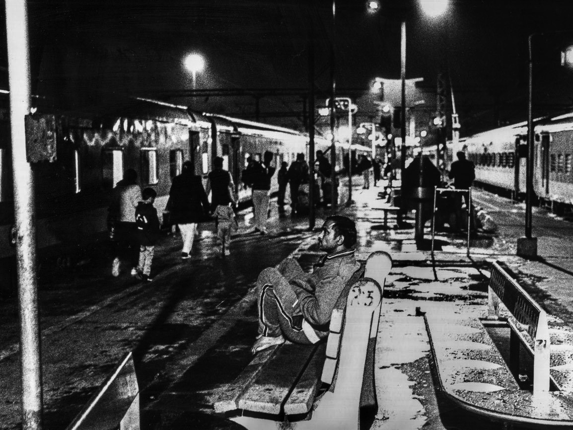 A man sits alone on a station bench at night, legs pulled up, watching passengers walk along a wet, lamplit platform beside parked trains