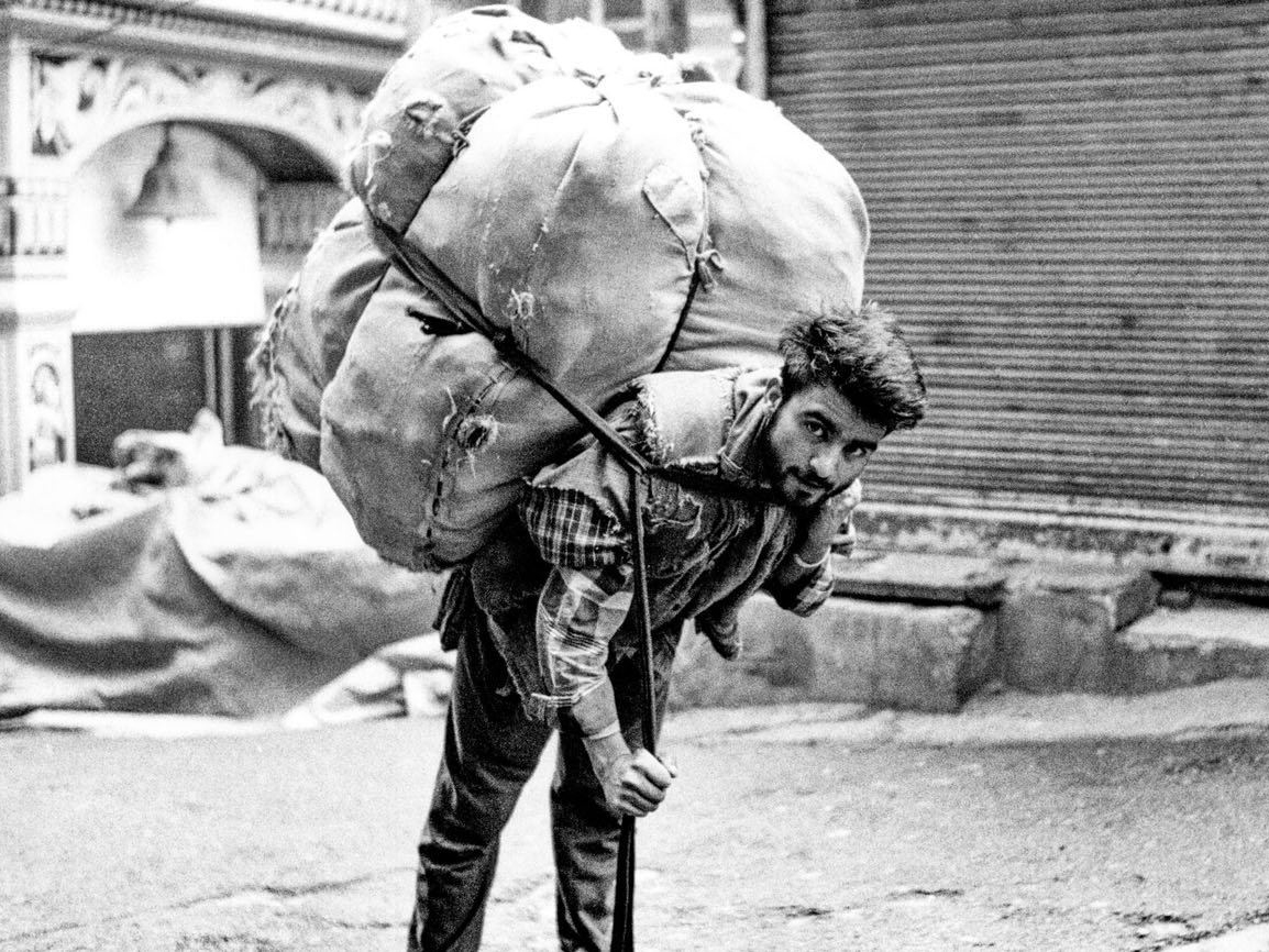 A man carries an enormous bundle strapped to his back and forehead with a rope, walking down a street