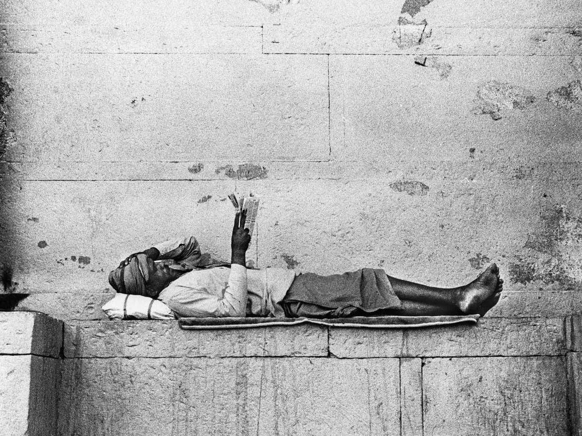 A man lies on a mat on stone steps, reading a folded newspaper held above his face, one bare foot resting against the worn stone