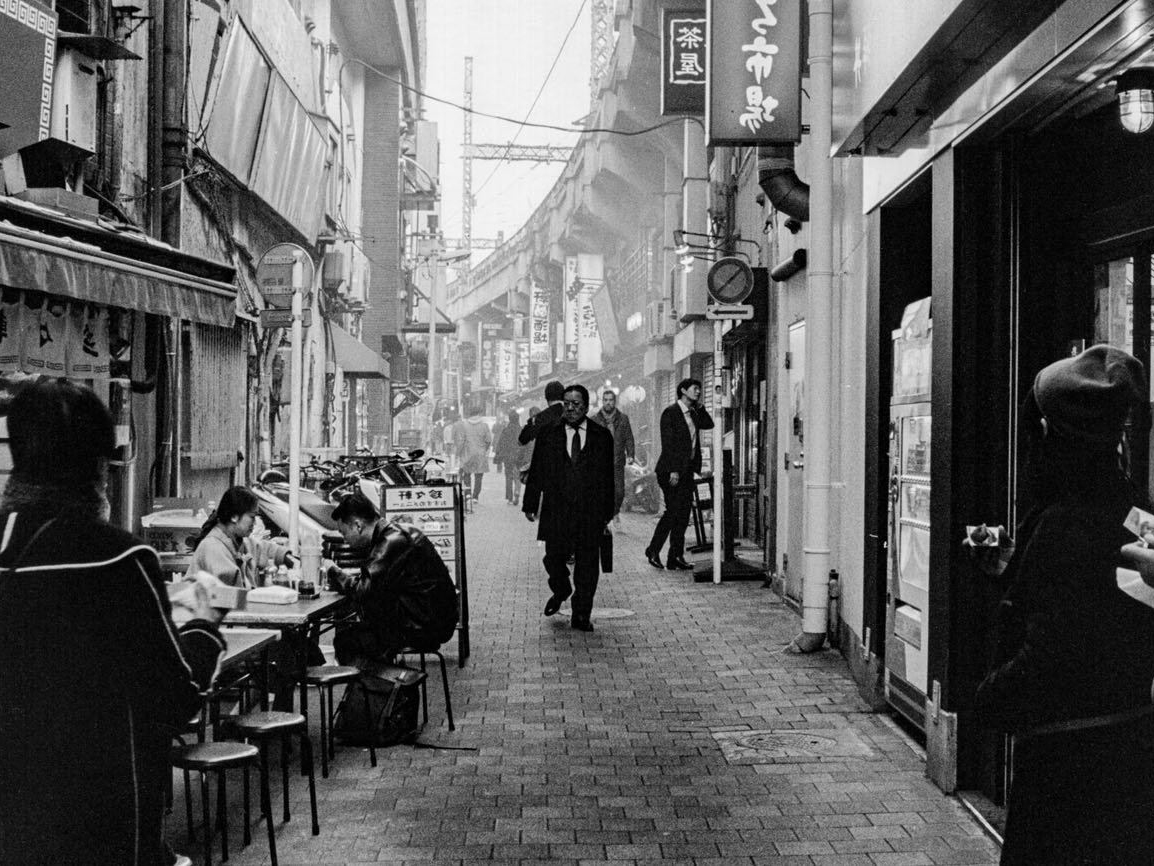 A man in a suit walks down a narrow paved alley lined with shops and hanging signs, past a small outdoor food stall where two people sit at a counter