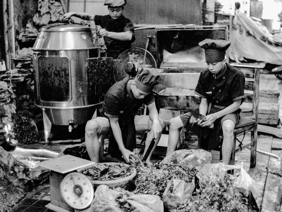 Two boys in chef hats and uniforms sort greens and vegetables on a stone ledge beside a large metal cooking drum and a hanging scale, on a busy street