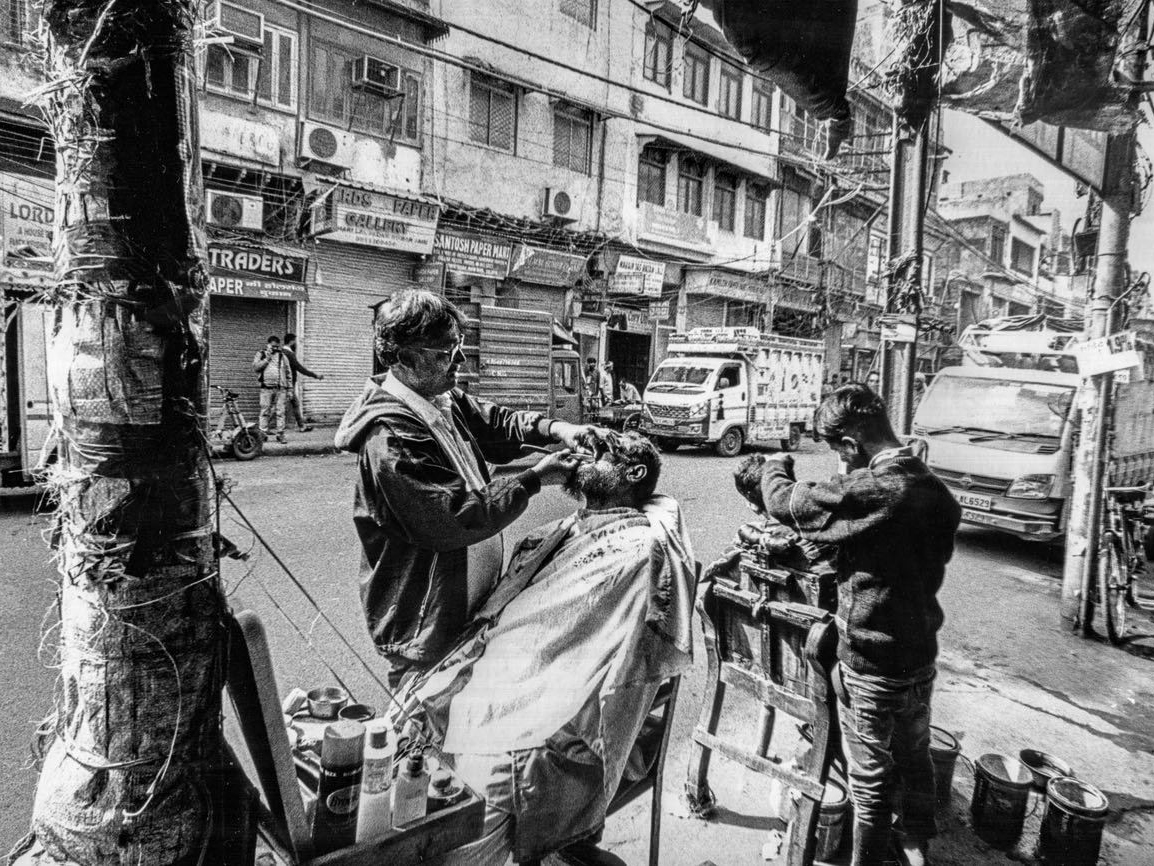 A street barber cuts a seated customer's hair at an open-air stall while a boy waits in a chair behind him, city traffic and shopfronts filling the background