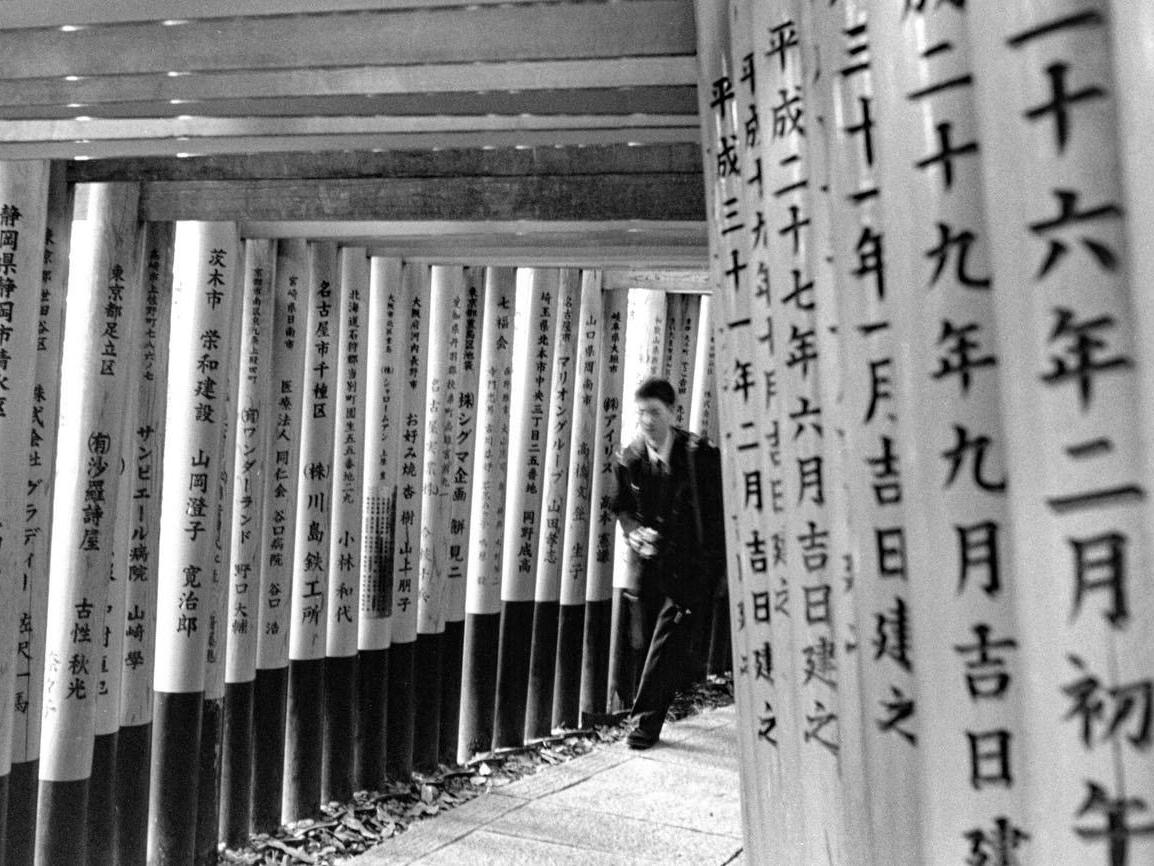 A man walks through a corridor of wooden torii gates densely inscribed with Japanese text, dappled light falling through the structure