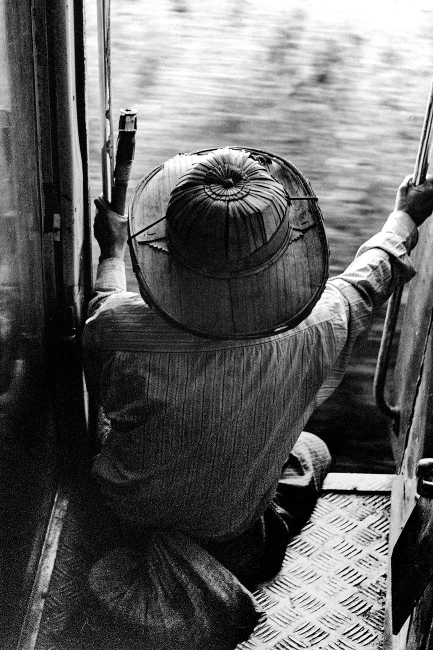 A person wearing a woven conical hat sits in the open doorway of a train carriage, one arm braced against the frame, motion-blurred landscape rushing past outside