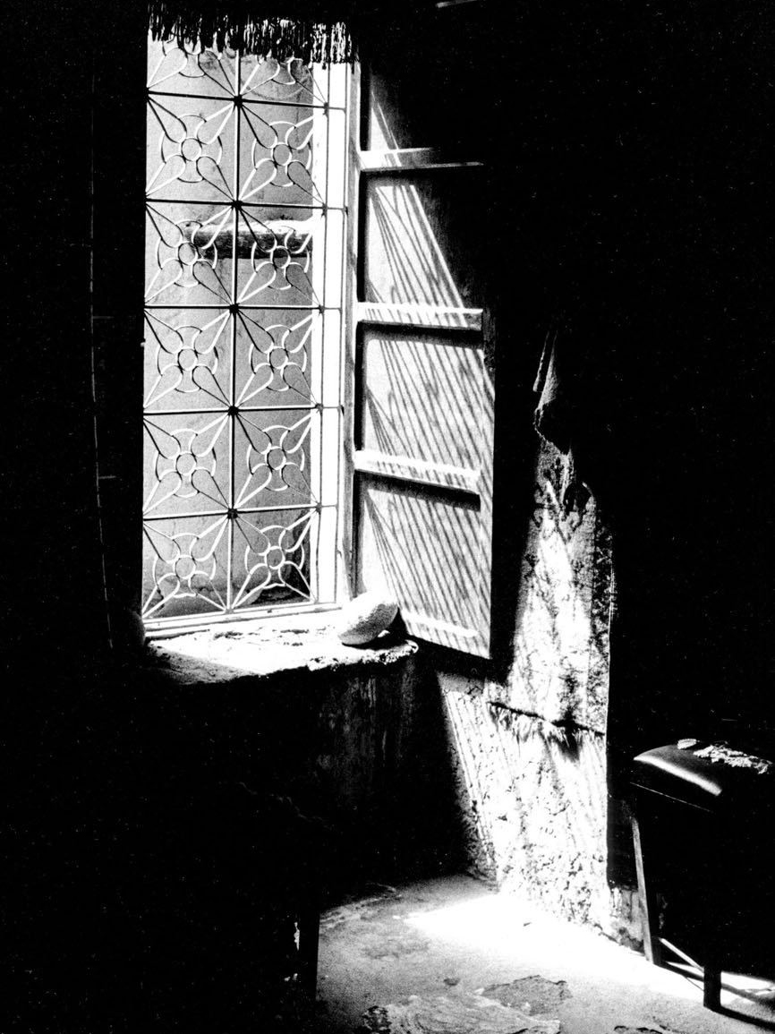 A shaft of light falls through a patterned window grille into a dark, textured stone interior, casting a striped shadow across the wall and floor