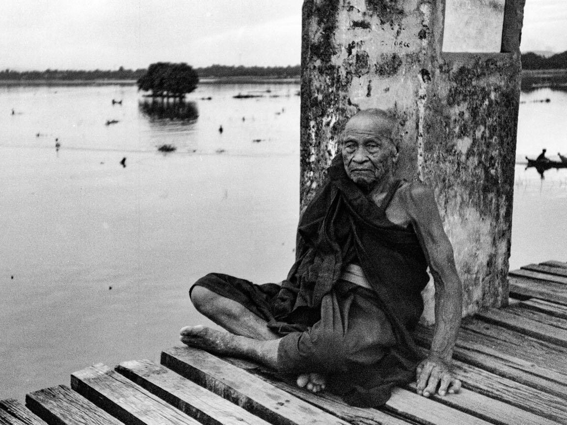 An elderly woman sits cross-legged at the edge of a wooden dock beside a stone pillar, looking toward the camera with a wide expanse of still water behind her