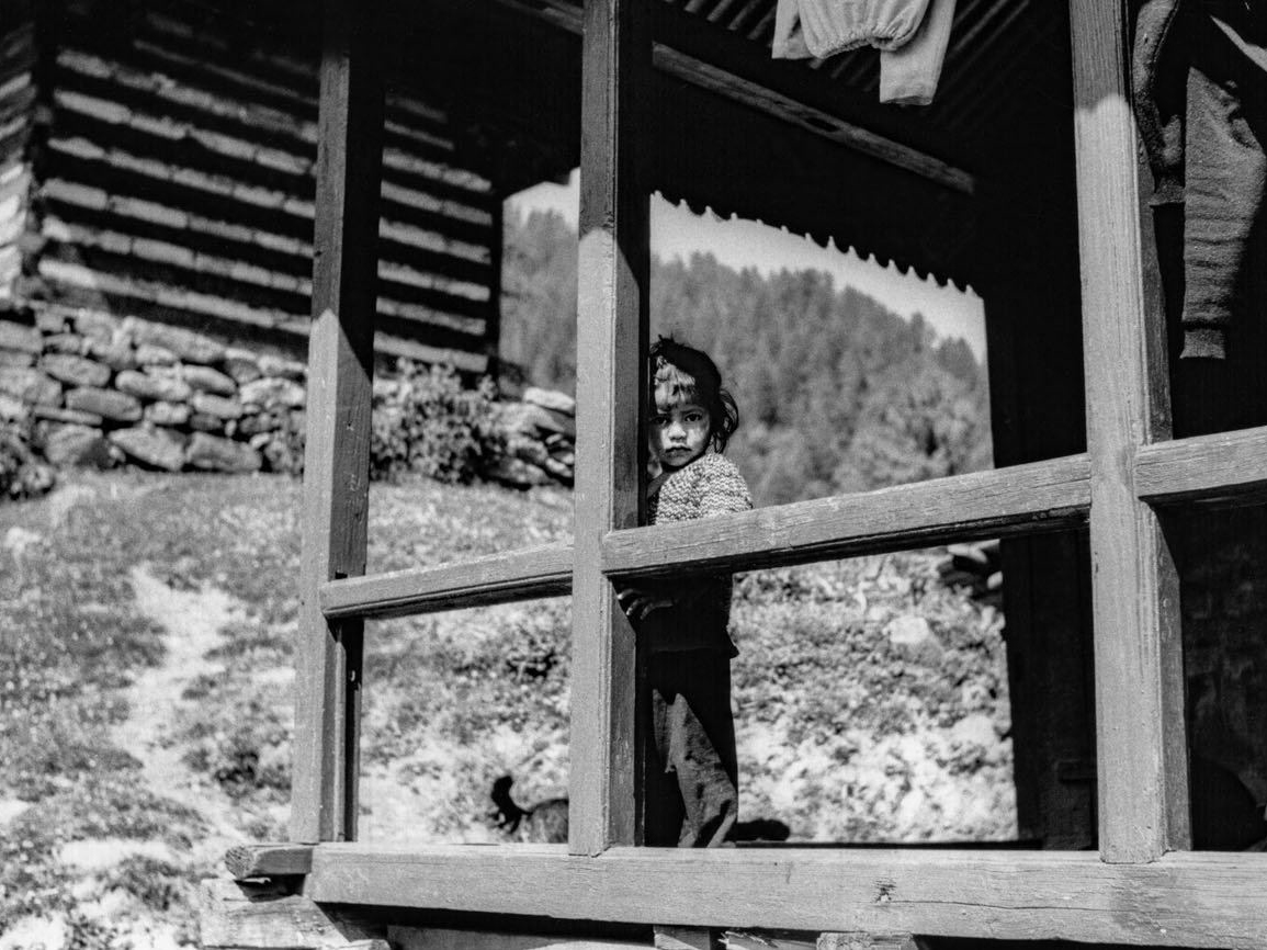 A young child peers around a wooden porch post on a mountain cabin, framed by railings and beams with a forested hillside in the background