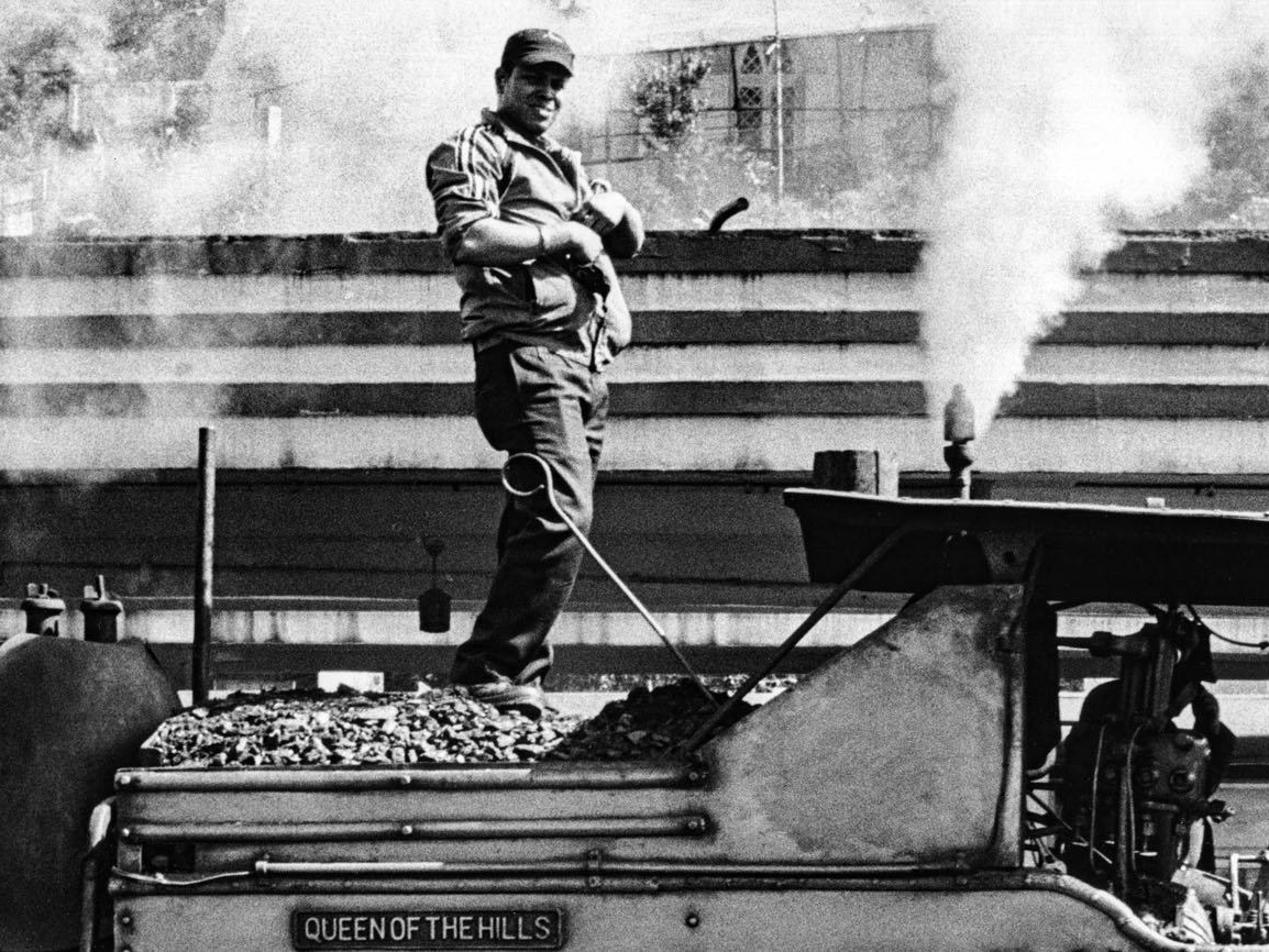 A man stands smiling atop a coal tender behind a labeled steam locomotive reading Queen of the Hills, steam rising around him