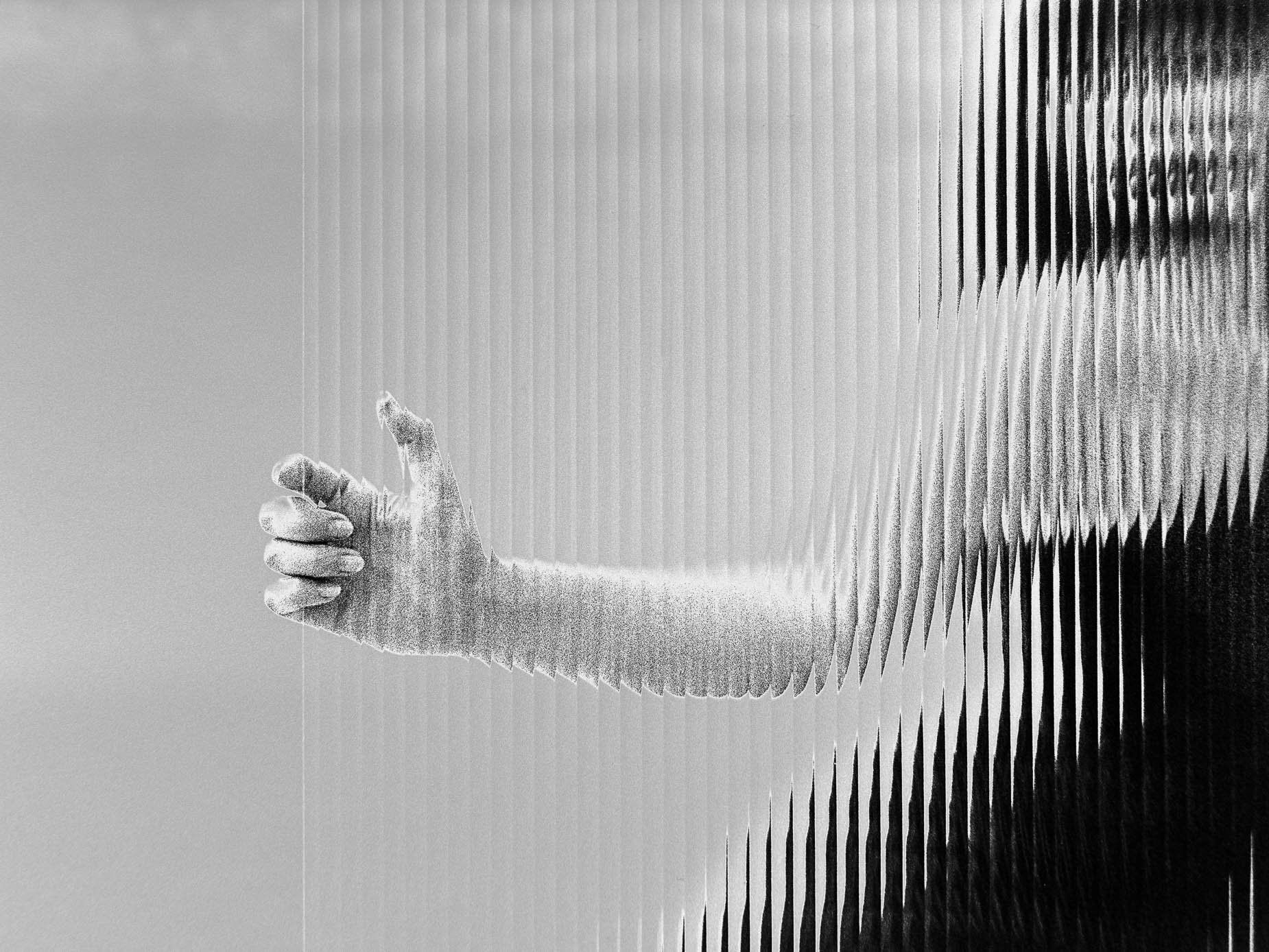 A hand and forearm cross a diagonal band of light and dark, dissolving into vertical striped bands as if seen through fluted glass