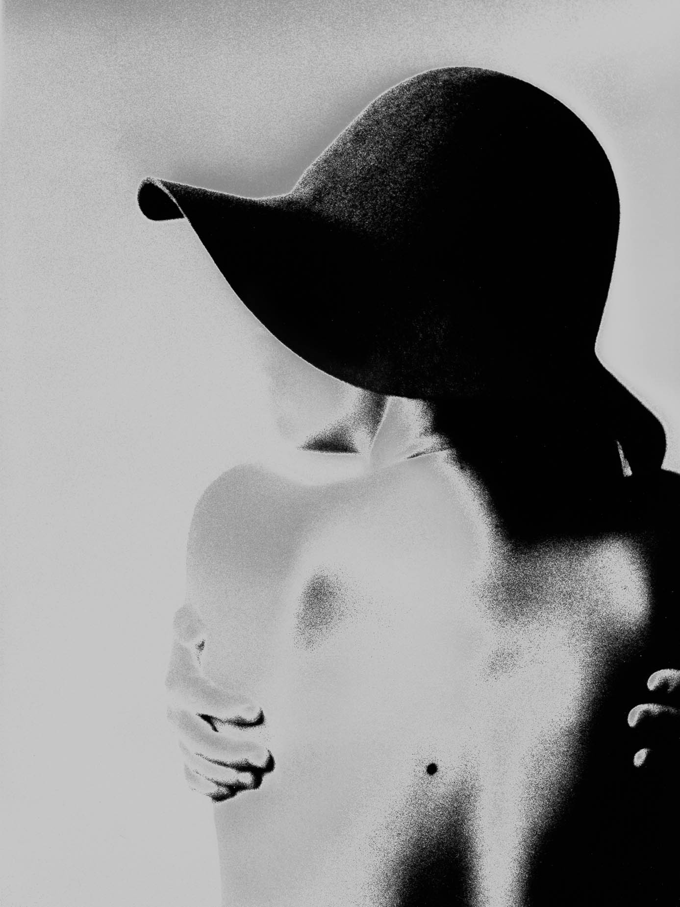 A wide-brimmed dark hat obscures a bare-shouldered figure's face entirely, one hand resting across the chest, solarised high-contrast lighting against a plain background