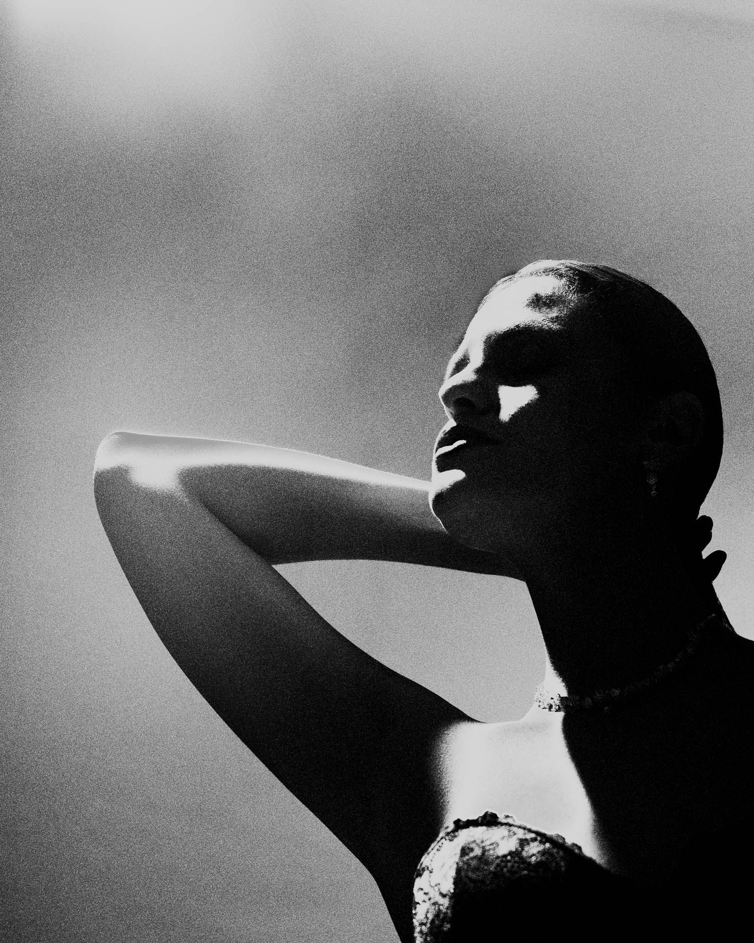 A figure with head tilted back and eyes closed, one hand raised to the throat, wearing a beaded necklace and matching earring against a dark background