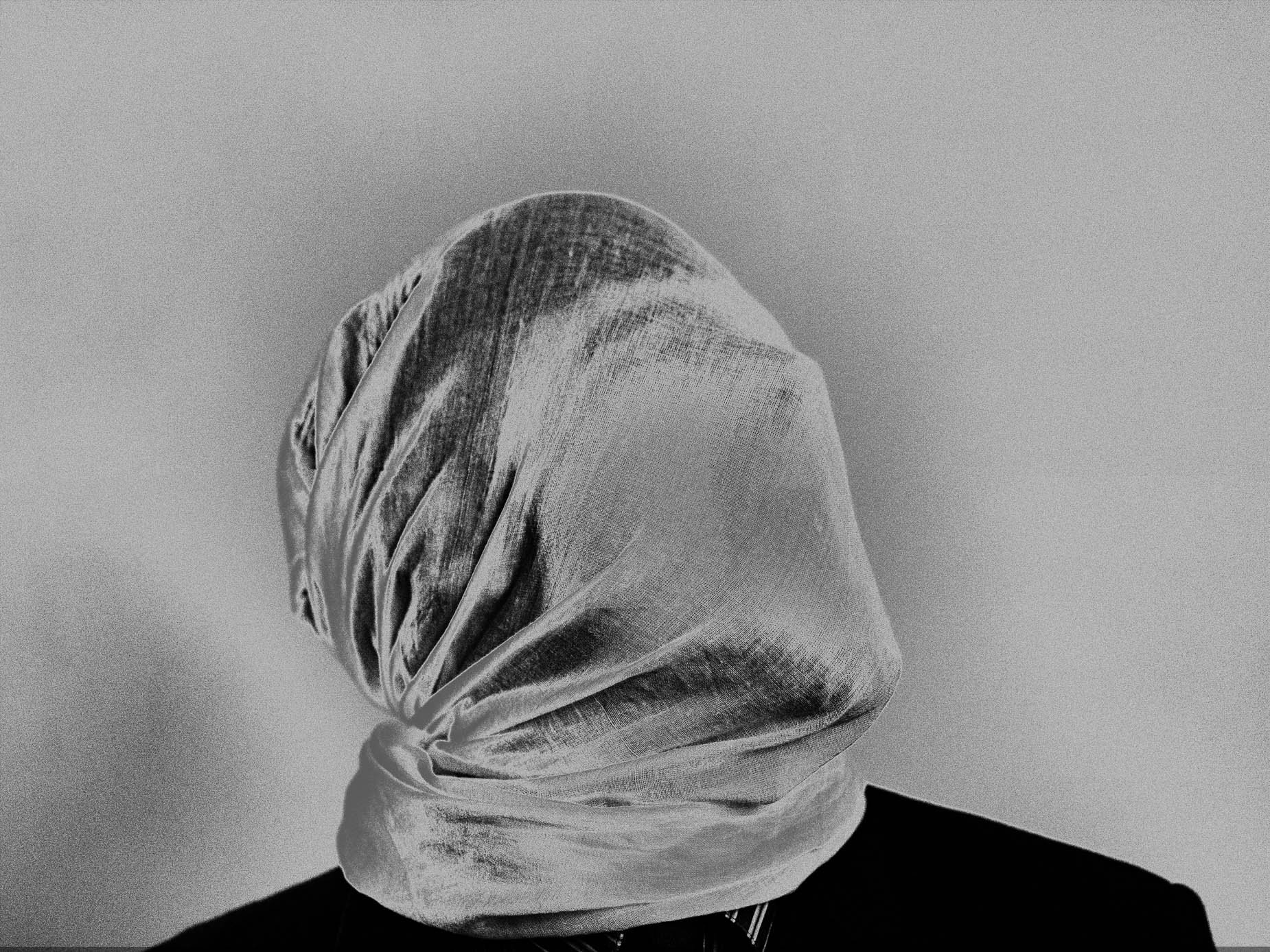 A head entirely wrapped and knotted in pale fabric, the face completely hidden beneath the twisted cloth, dark jacket visible at the shoulders