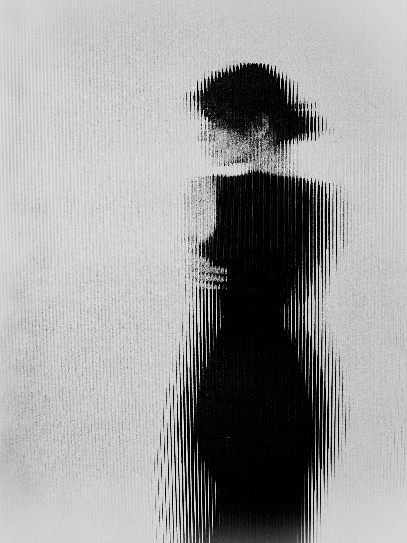 A veiled silhouette behind fluted glass, the head crowned by a dark rounded shape, the whole body broken into vertical bands against a pale background