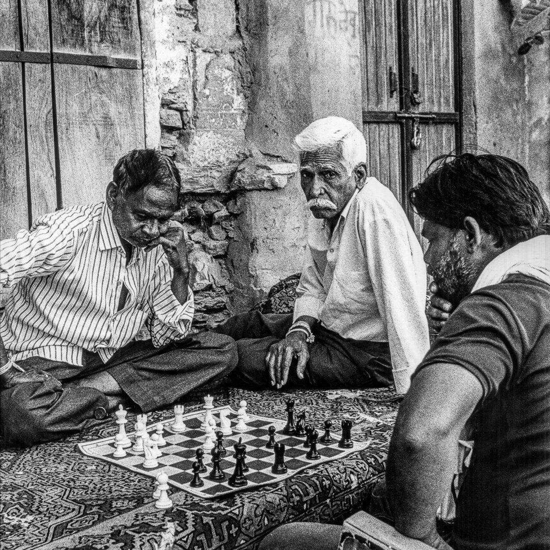 Three men sitting on a patterned rug playing chess in a weathered stone alleyway, one with white hair and a moustache looking directly at the camera, an old wooden door behind them