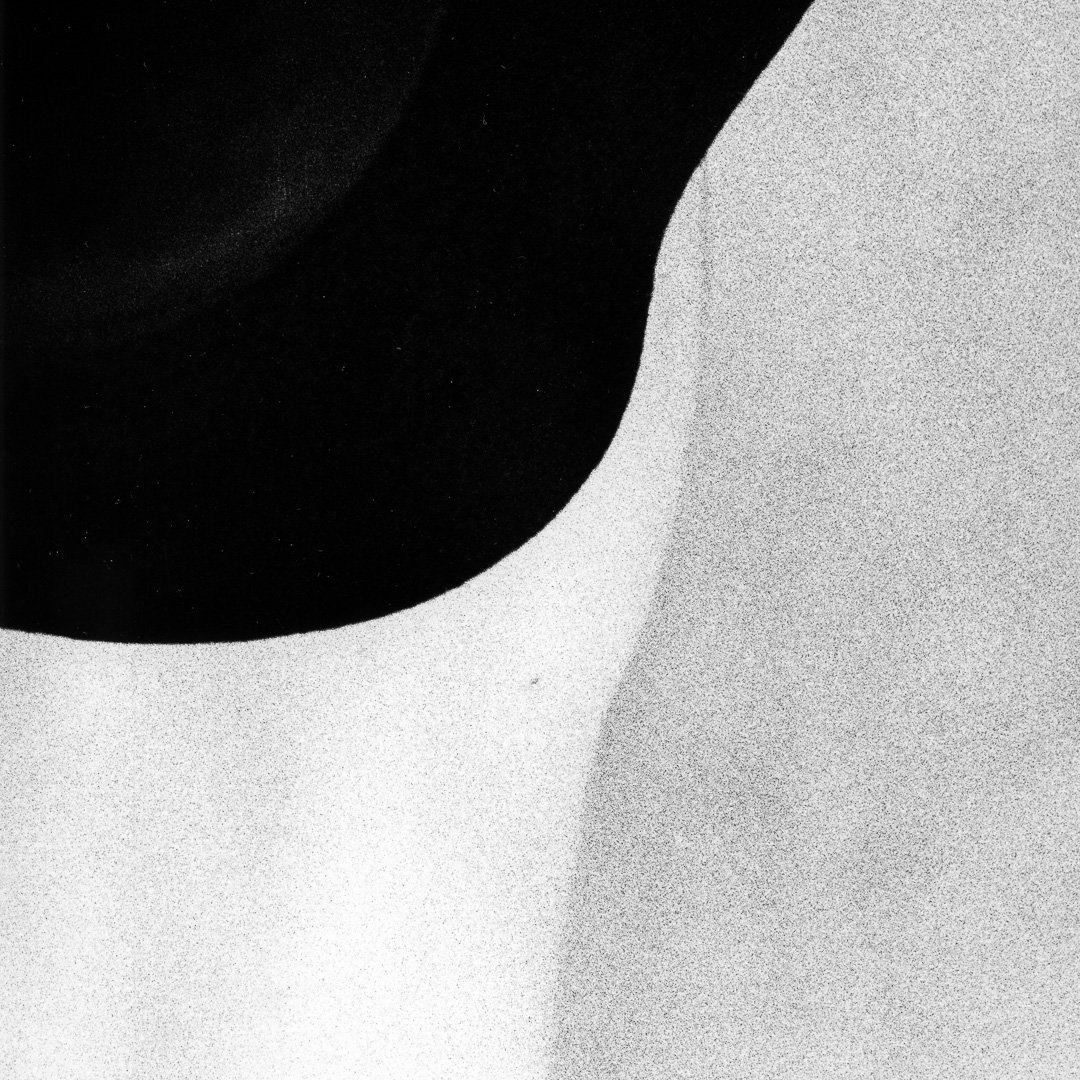 Abstract high-contrast composition where a dark curved hat brim meets a pale bare shoulder, forming two graphic shapes divided by a soft grainy edge