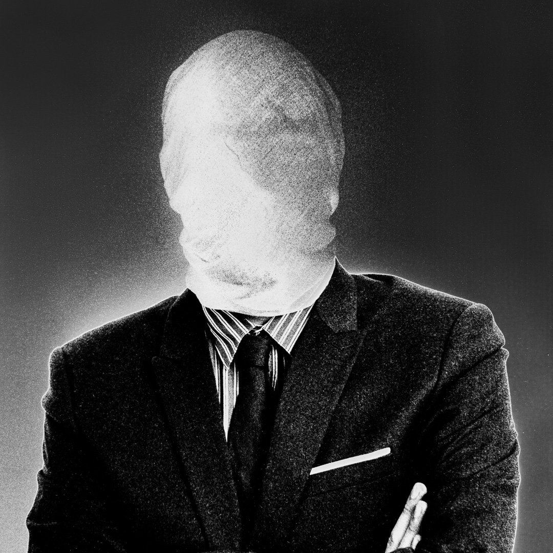 A man in a dark suit and striped tie, arms crossed, his head and face entirely wrapped in pale grainy fabric with no features visible