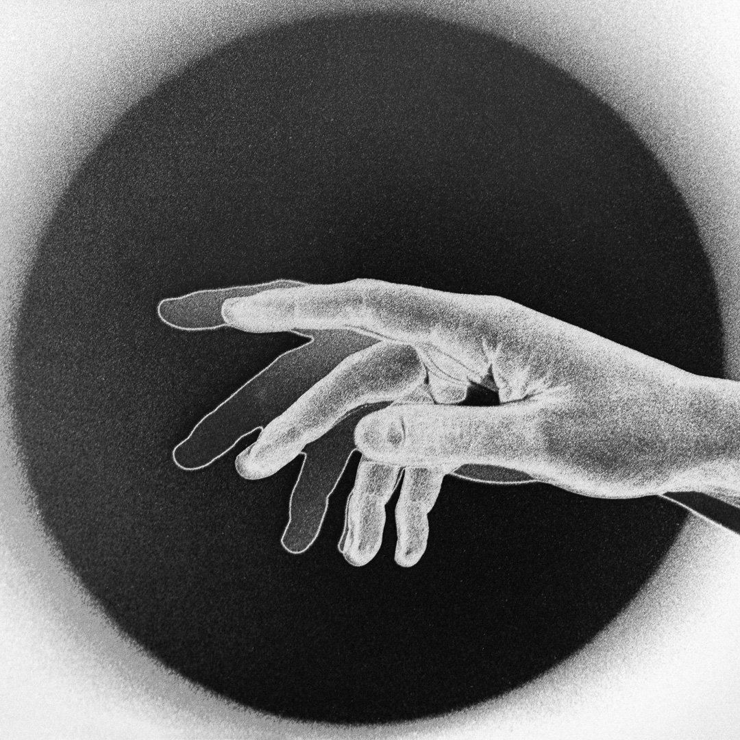 A hand with fingers curled reaching into a large dark circular void against a pale background, silver gelatin grain visible throughout