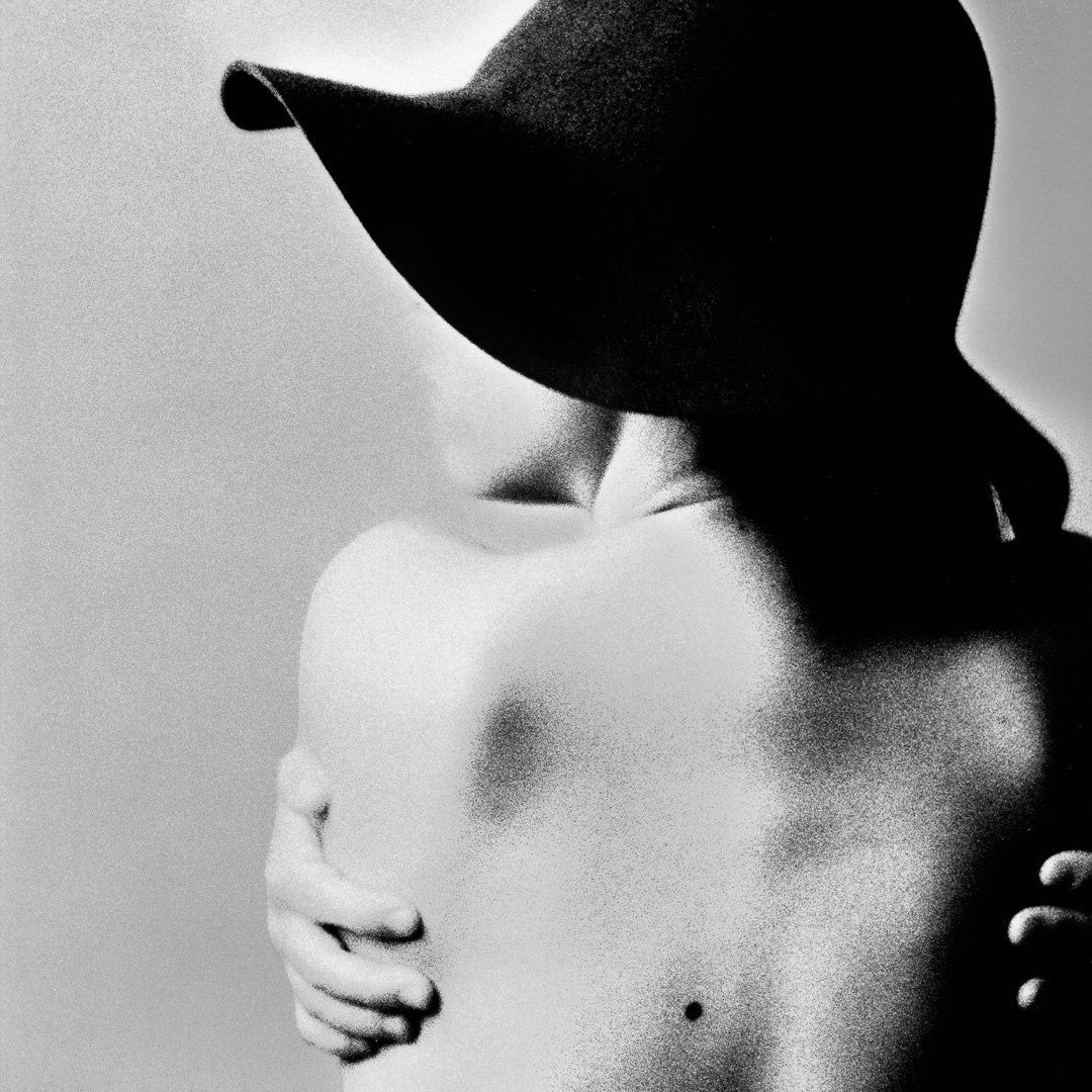 Close crop of a bare shoulder and neck, the face hidden beneath a wide black hat brim, one hand resting across the chest gripping the opposite arm